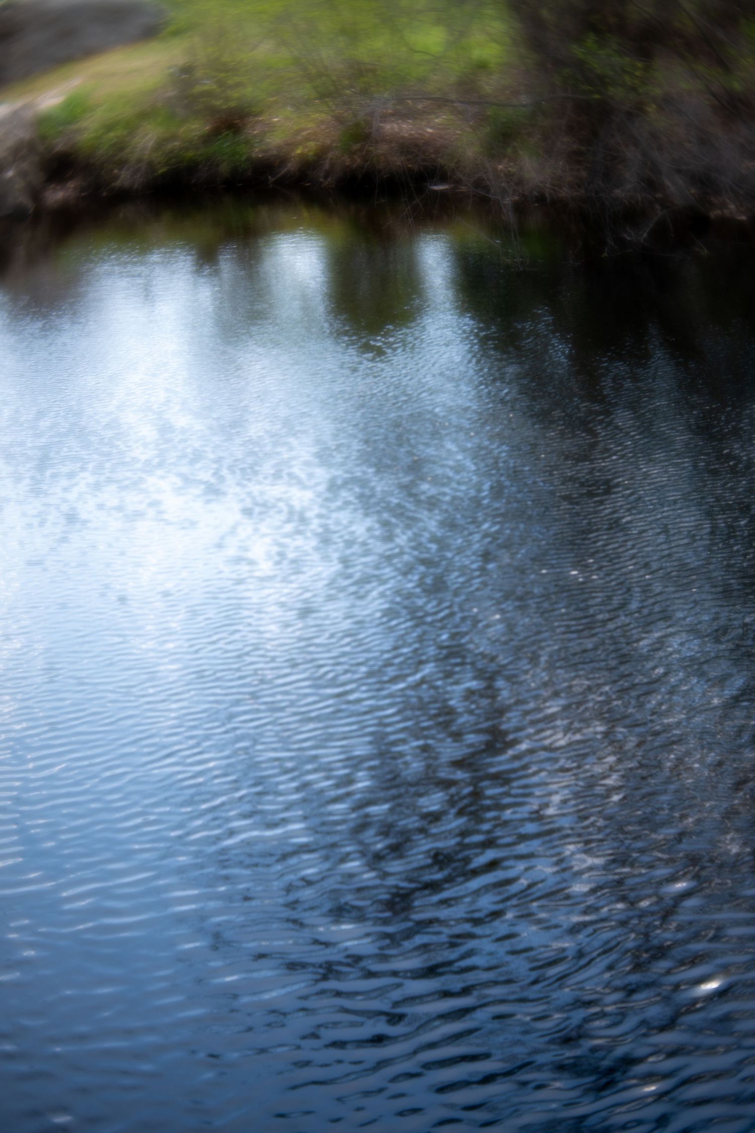 Blurry artistic photo of a calm blue river with rippling water reflecting trees and green banks on a cloudy day.