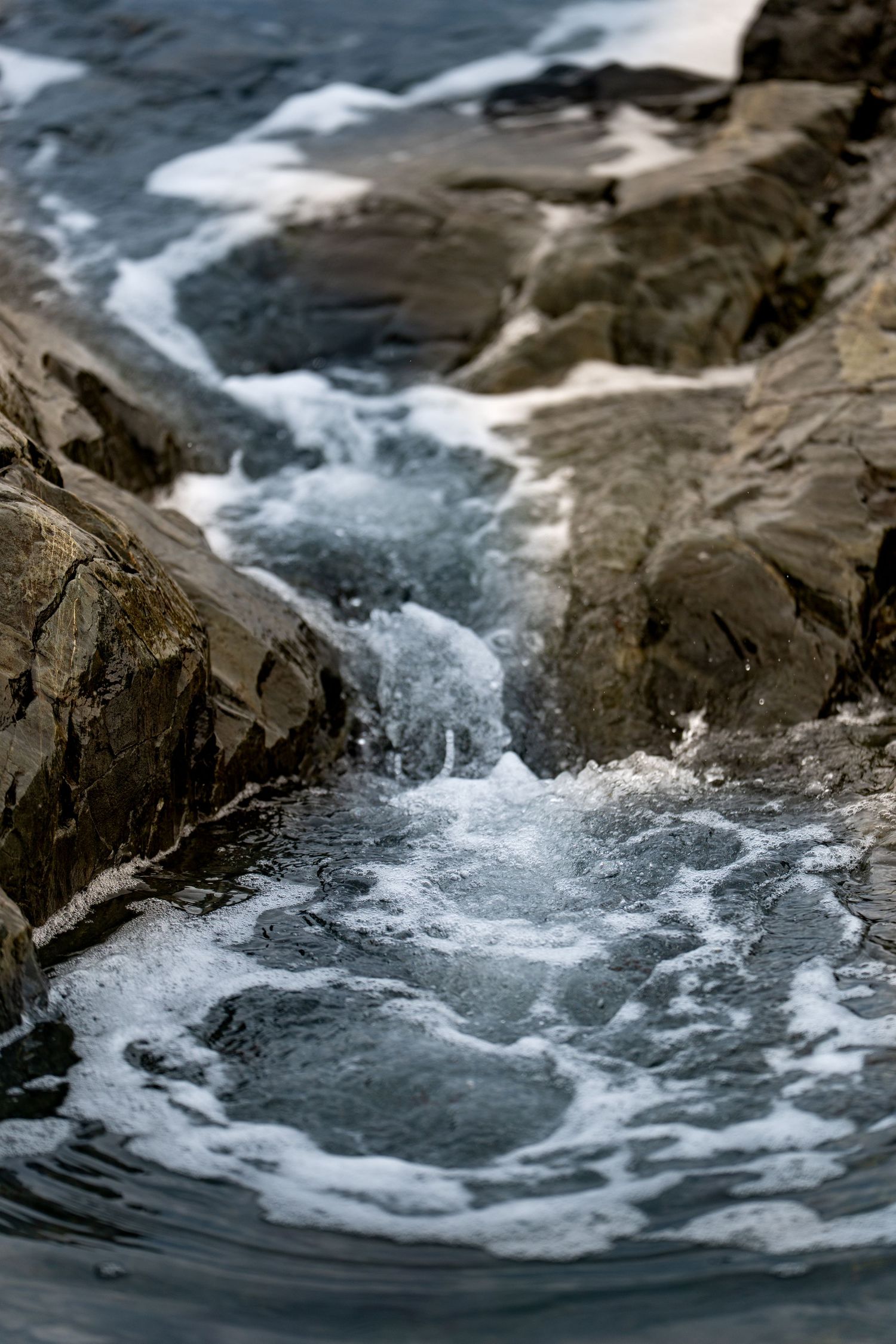 Ocean waves crash through a narrow rocky crevice, creating white sea foam and swirling water pools along the rugged coastline.