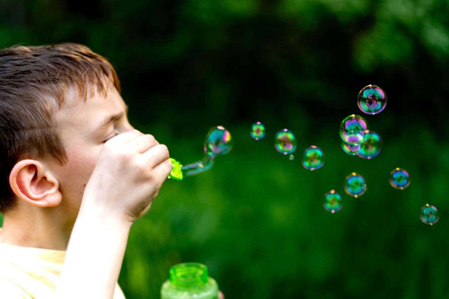 A young boy blowing colorful soap bubbles outdoors with green trees in the background on a sunny day.
