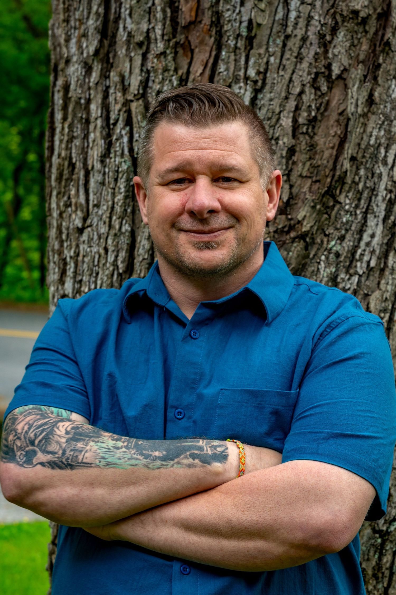Person wearing blue polo shirt stands confidently against large tree trunk background.