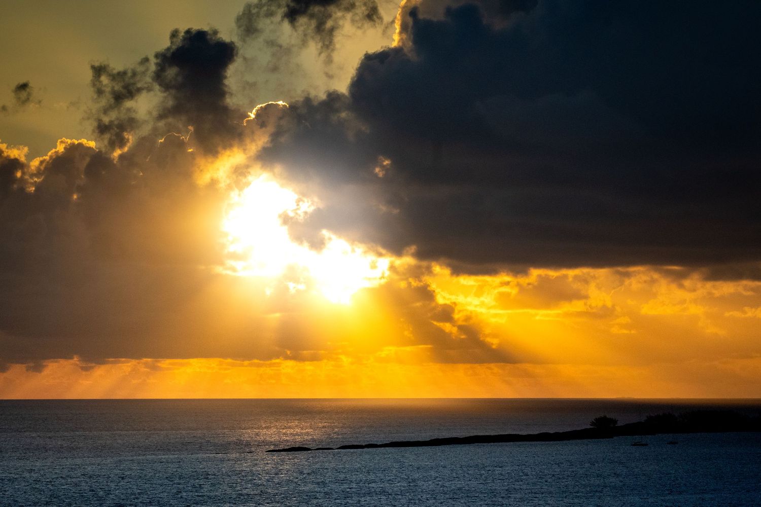 Dramatic sunset over ocean horizon with dark clouds and golden sunbeams breaking through creating silhouette effect.