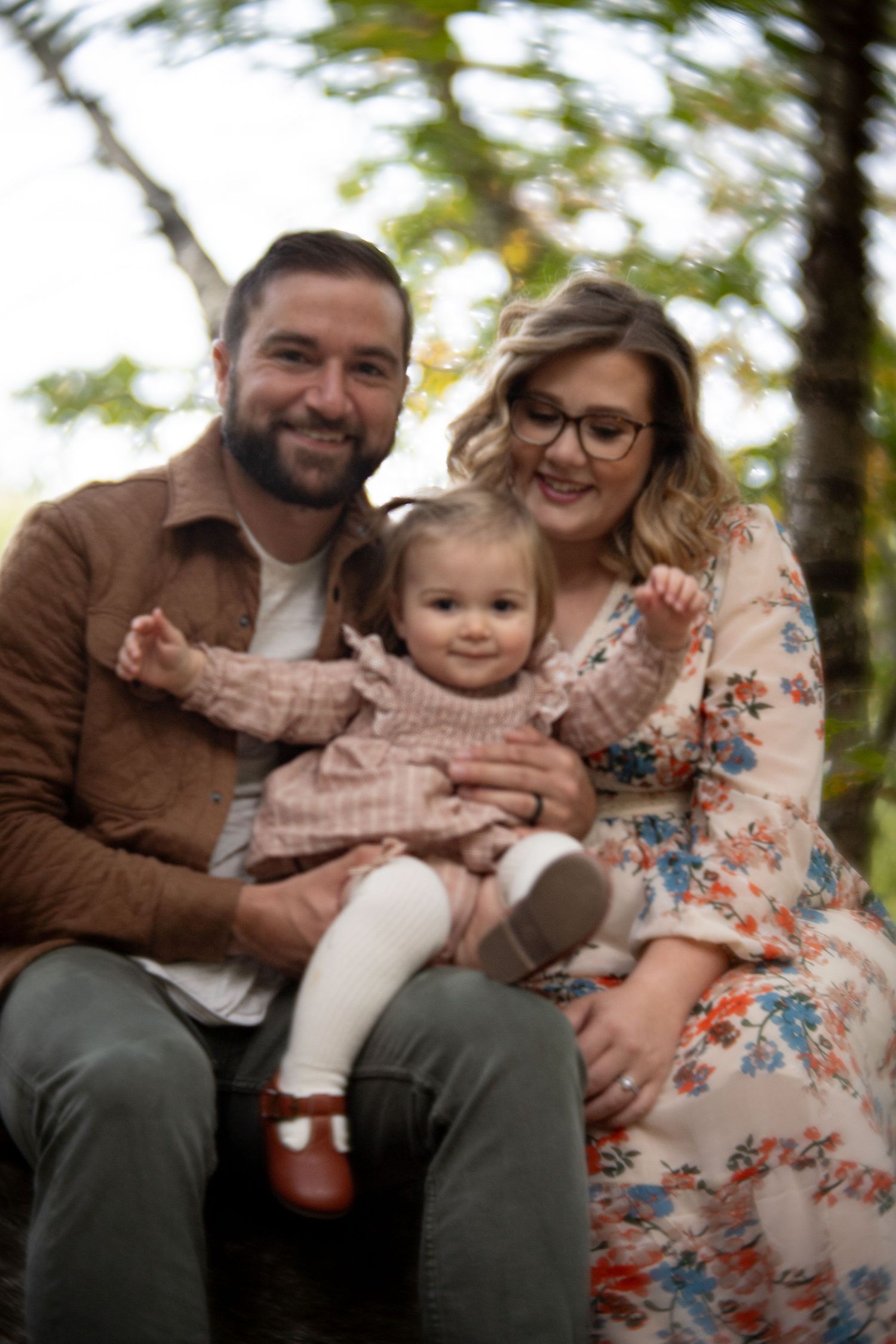 A family sits together outdoors in autumn, with a small child in pink between two adults.