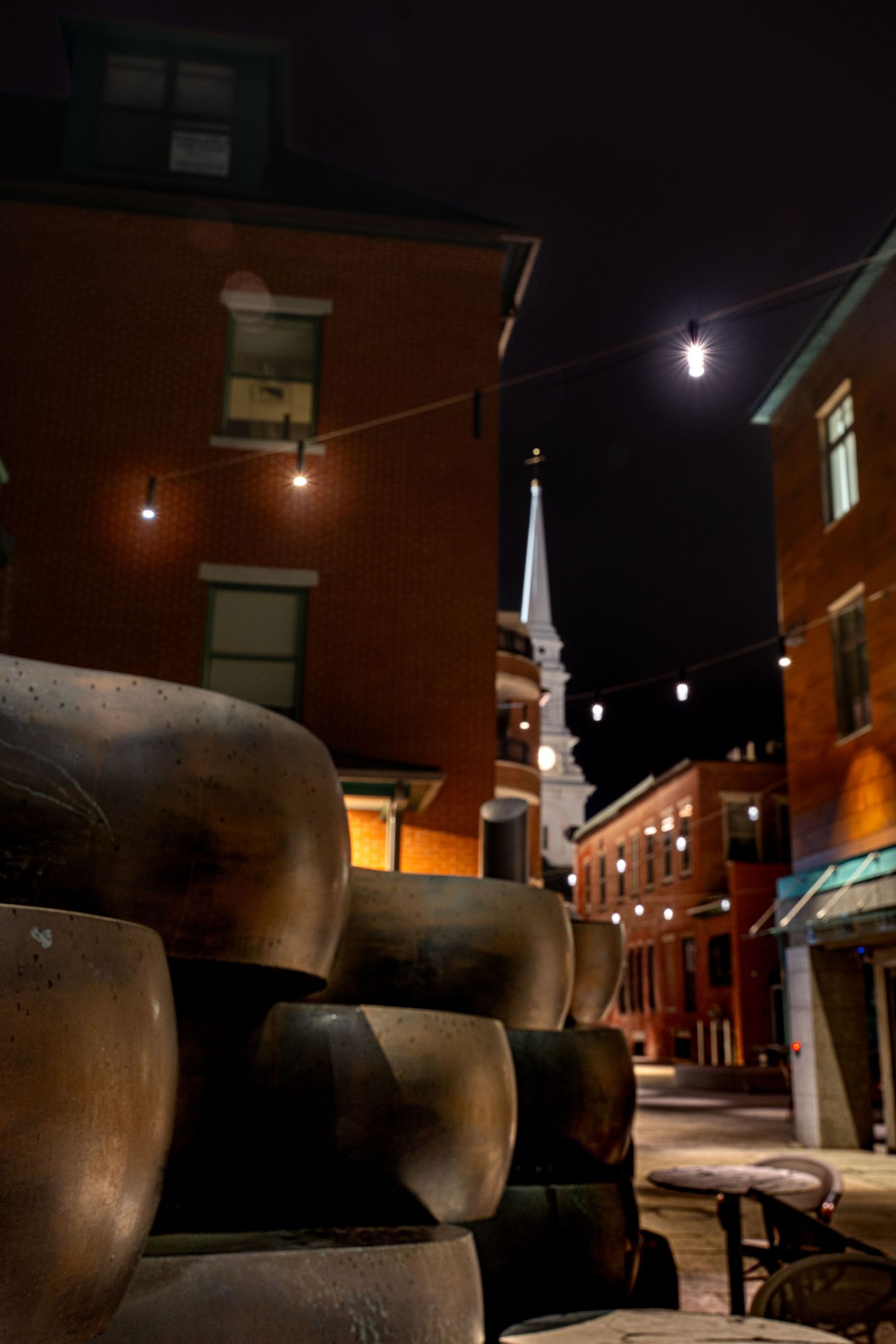 Historic brick buildings and church steeple lit at night with string lights and antique cannons in foreground.