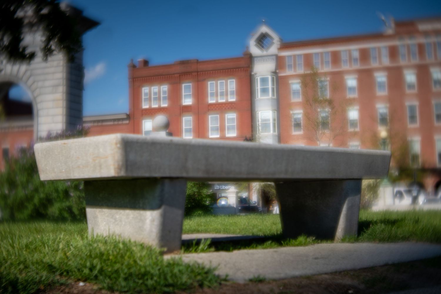 A concrete bench sits in a green urban park with a red brick building and clock tower in the background.