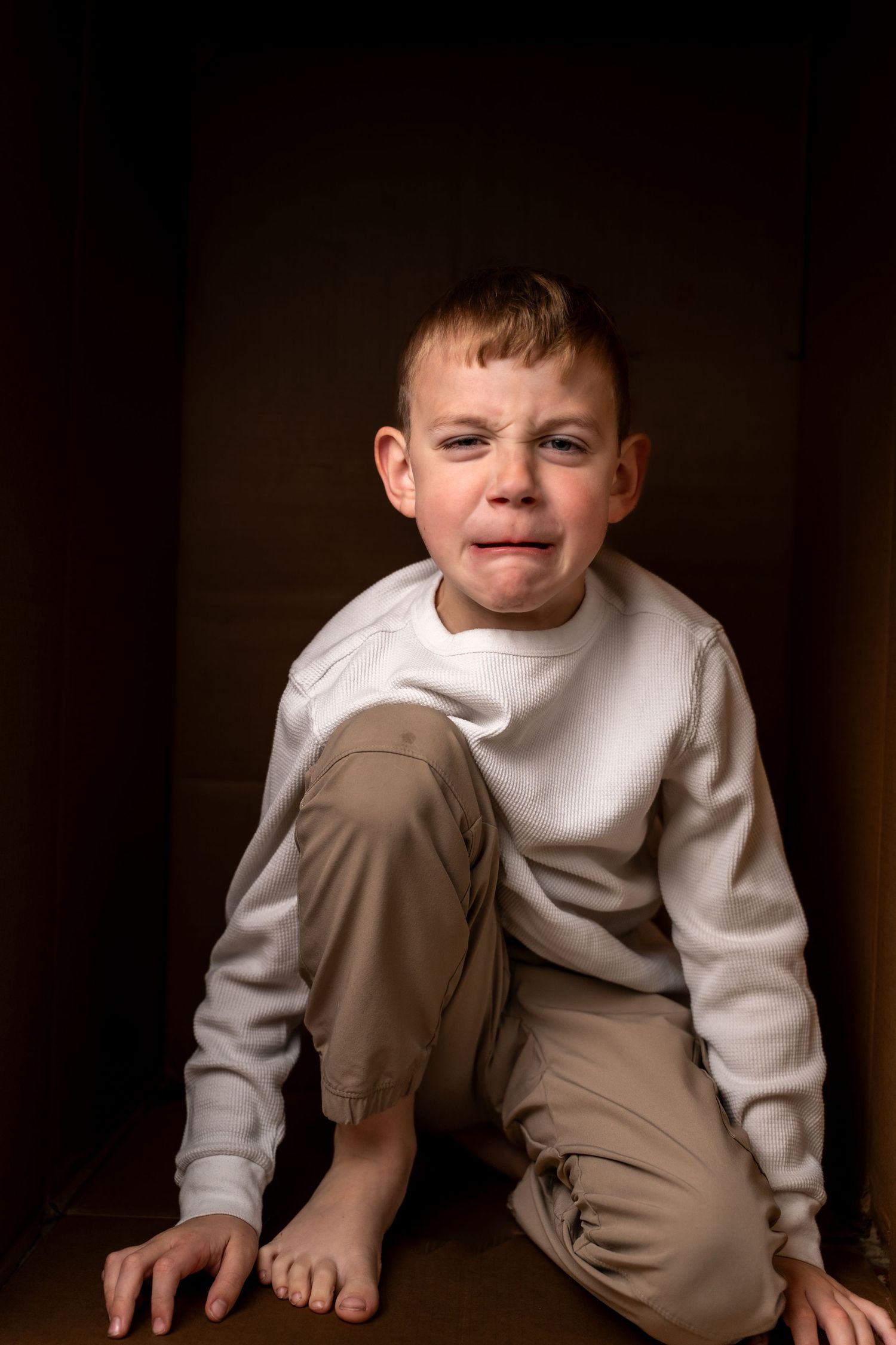 A young boy sitting barefoot in a dark corner, crying with a distressed expression, wearing a white sweatshirt and beige pants.
