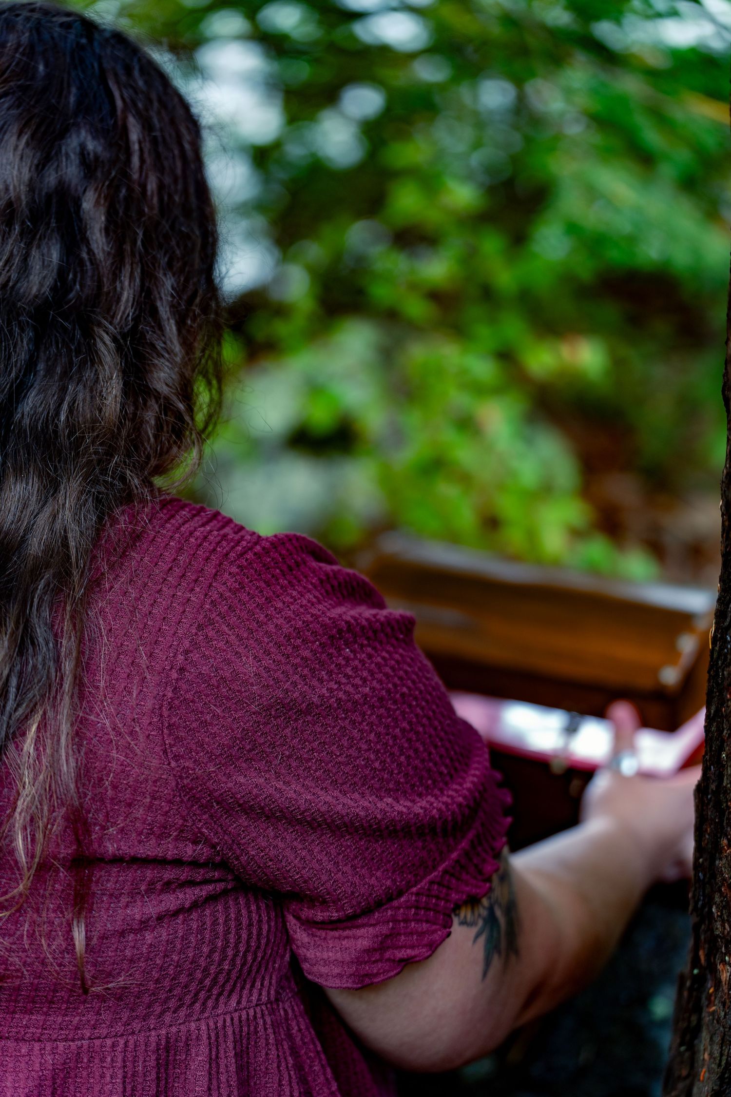 Person in a burgundy sweater sits at a window looking out at green foliage in soft focus.