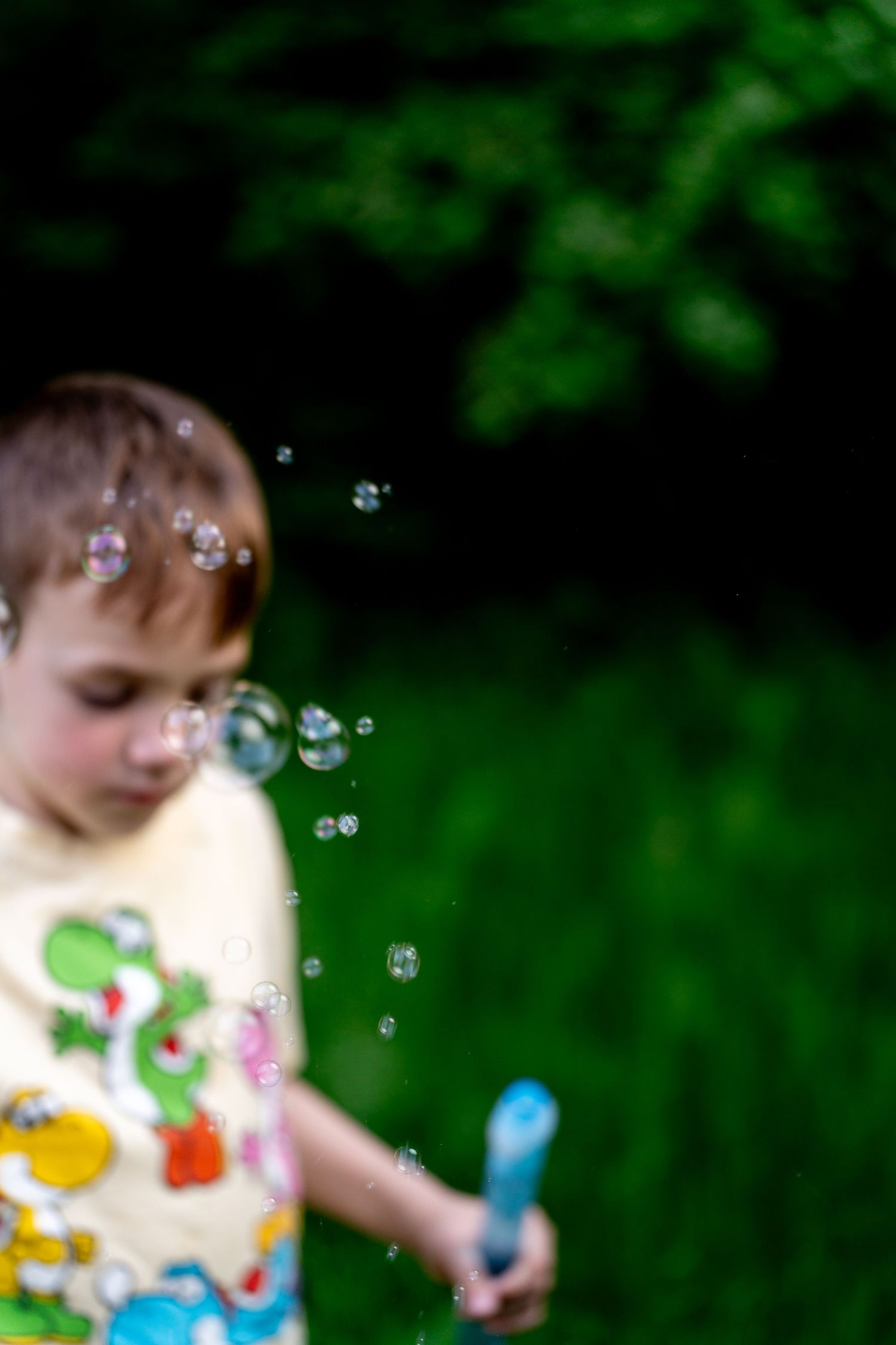 A young boy in a colorful Yoshi shirt blows soap bubbles outdoors with a blue bubble wand against a green background.