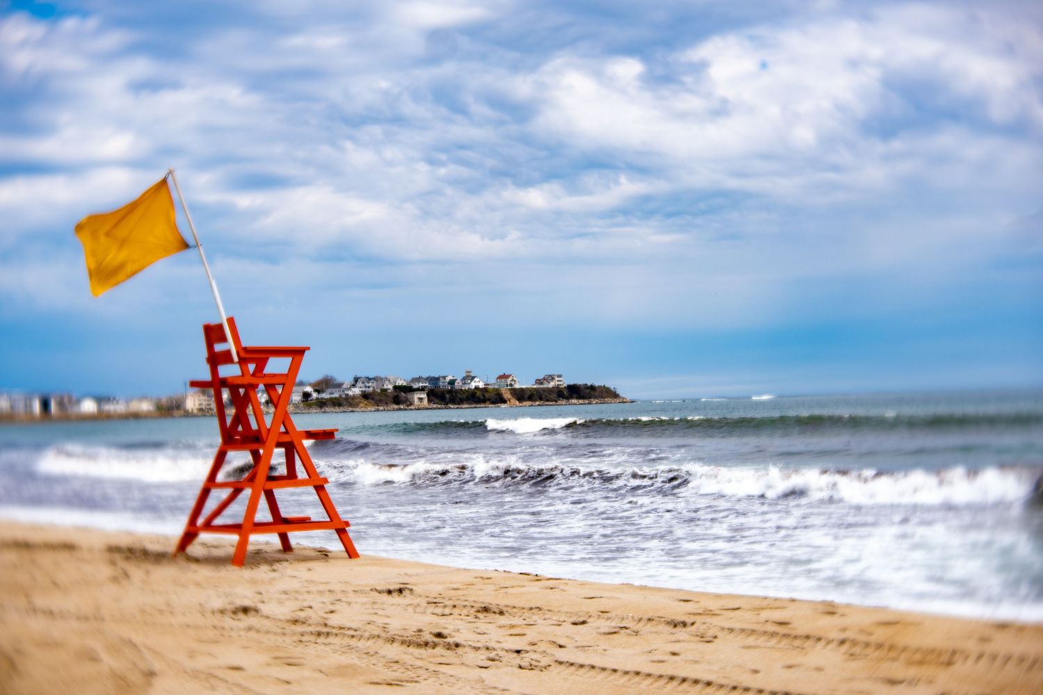 Red lifeguard chair with yellow flag on sandy beach beside ocean waves under cloudy blue sky.