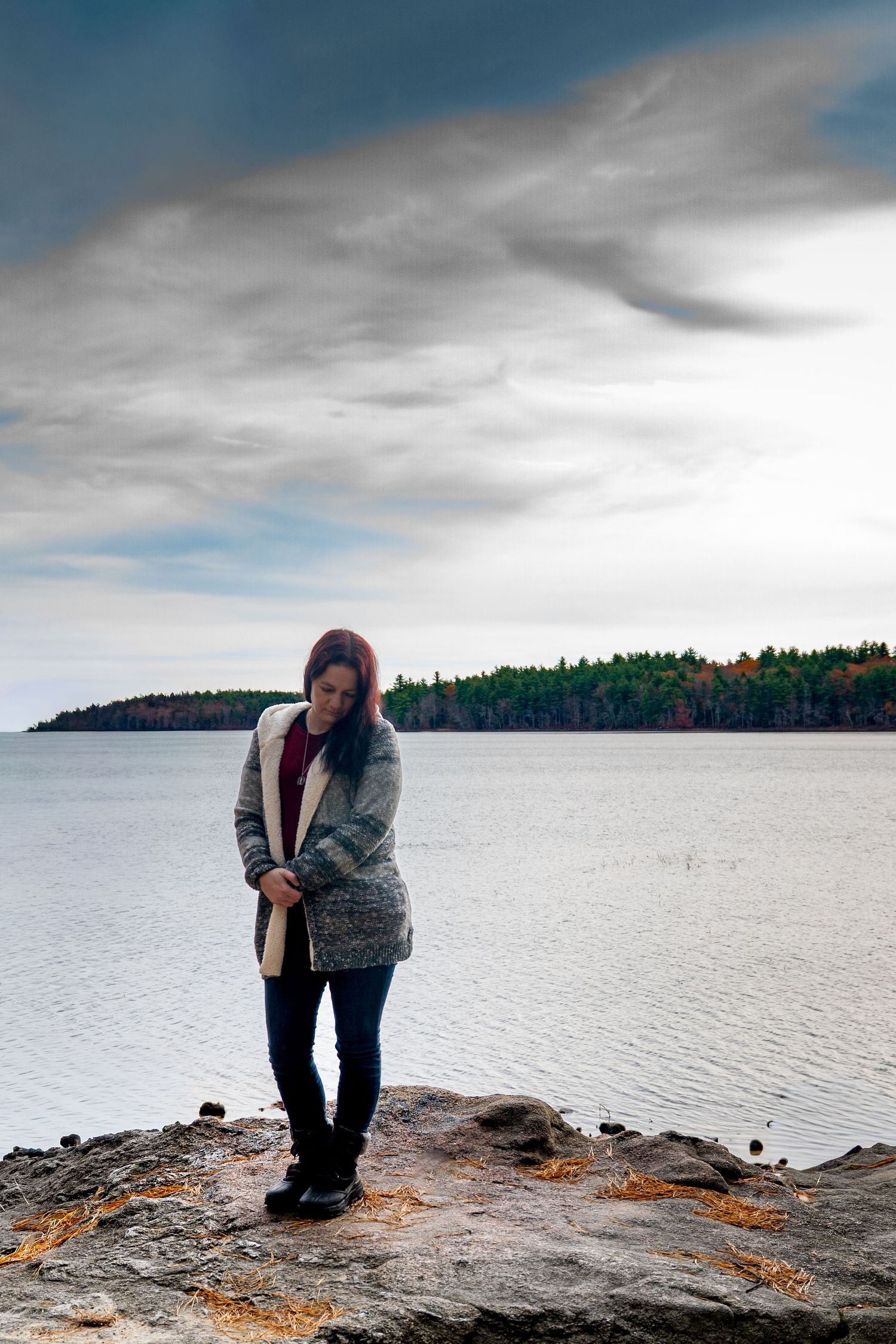 A person in a gray sweater stands on a rocky shoreline overlooking a misty lake with forested islands in the distance.