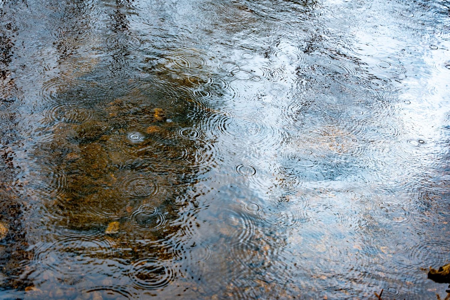 Rippling water surface with rain droplets creating circular patterns over rocky riverbed with reflections.