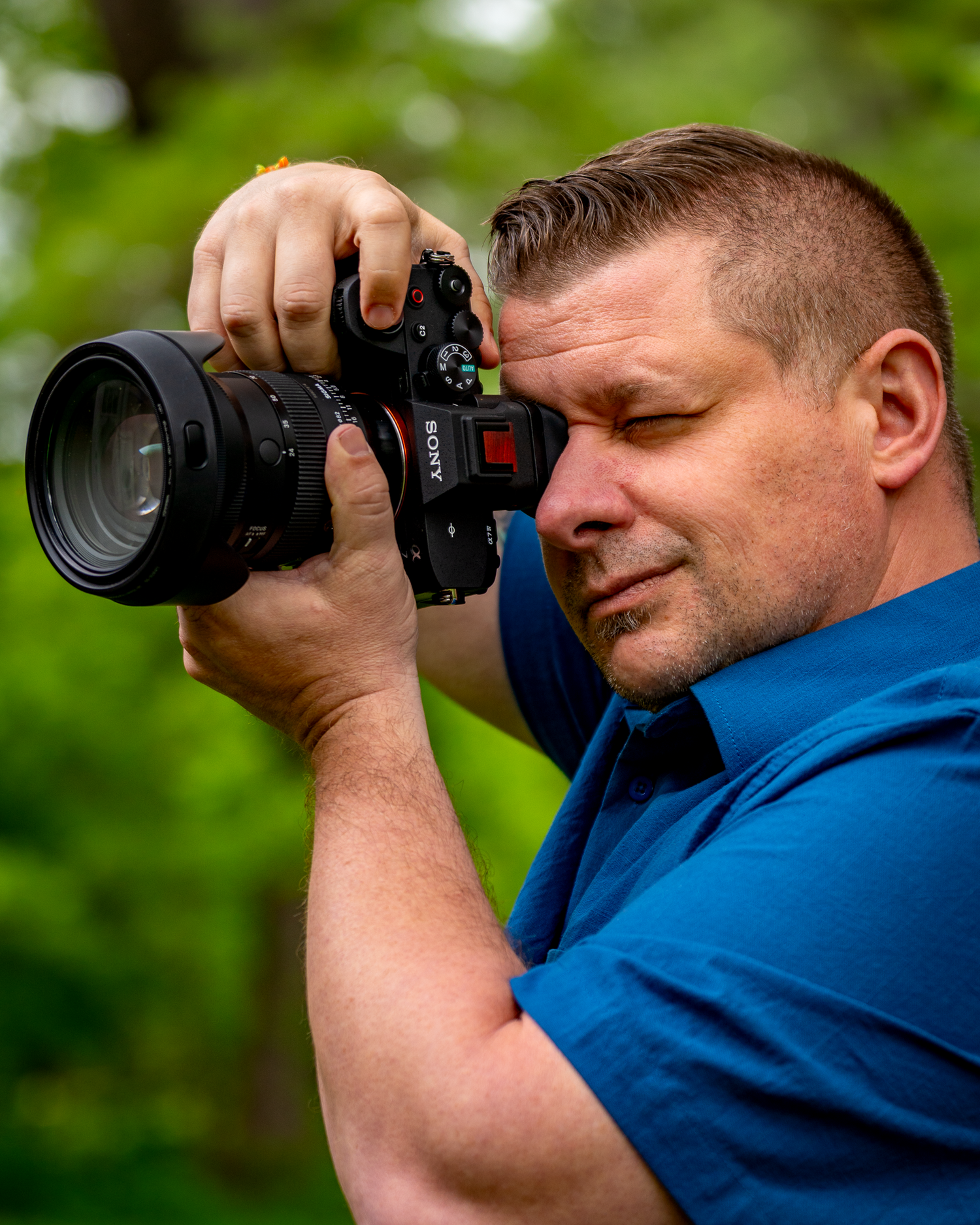 Photographer wearing blue shirt takes photos with professional DSLR camera outdoors.