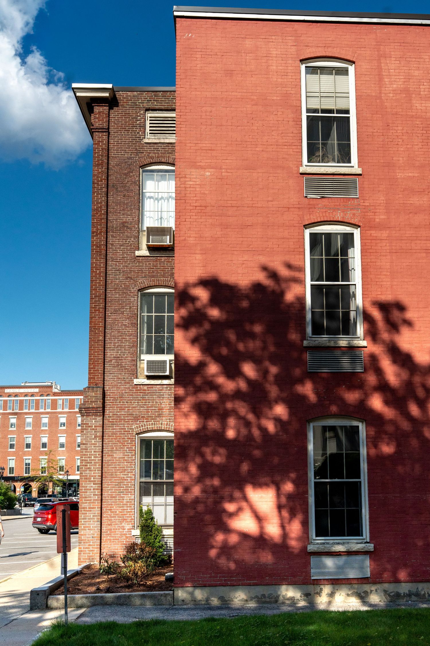 Historic red brick multi-story building with arched windows casting tree shadows on sunny day.