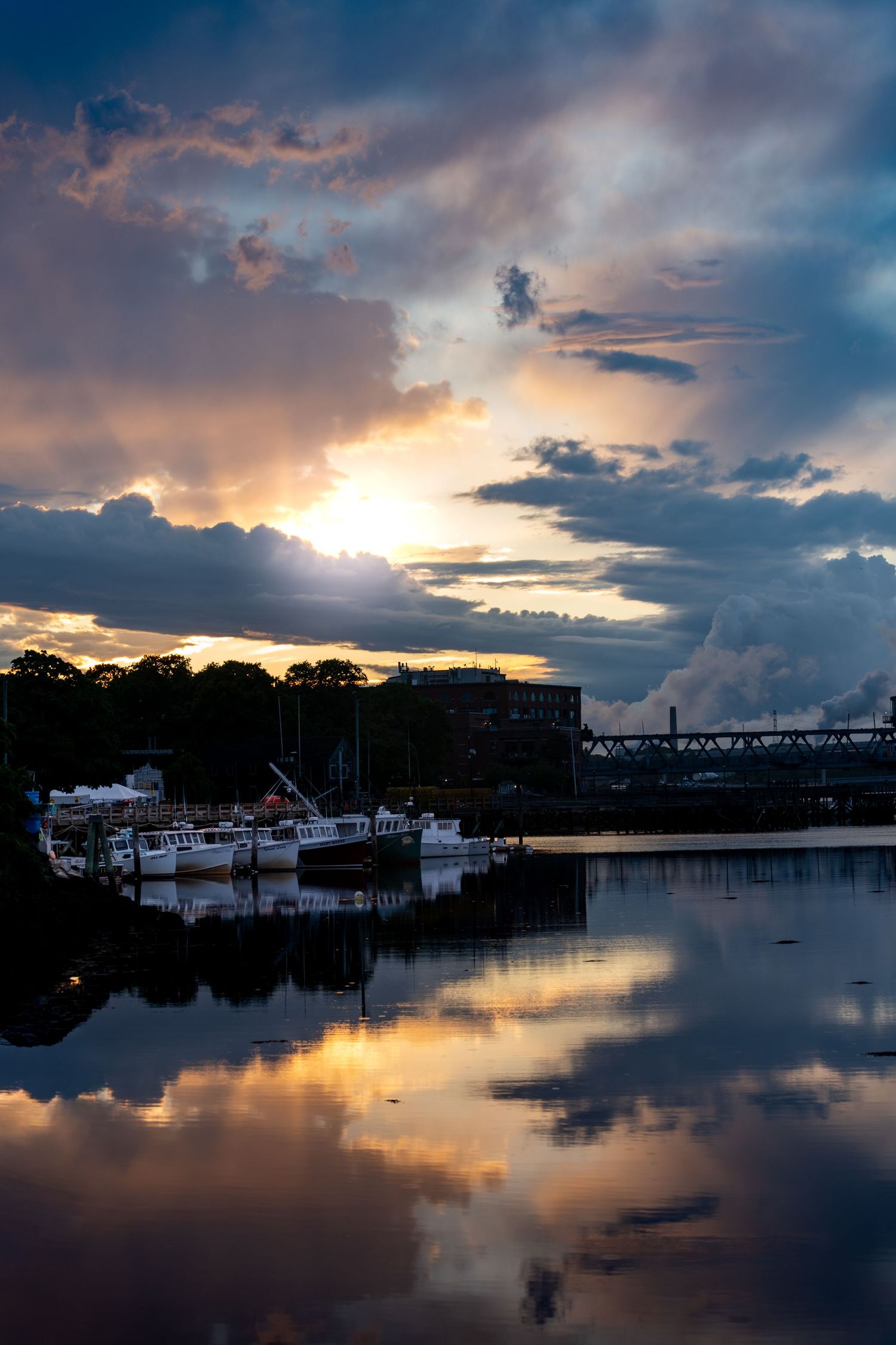 Stunning sunset over a calm harbor with boats docked, dramatic clouds reflected in still water near a bridge.