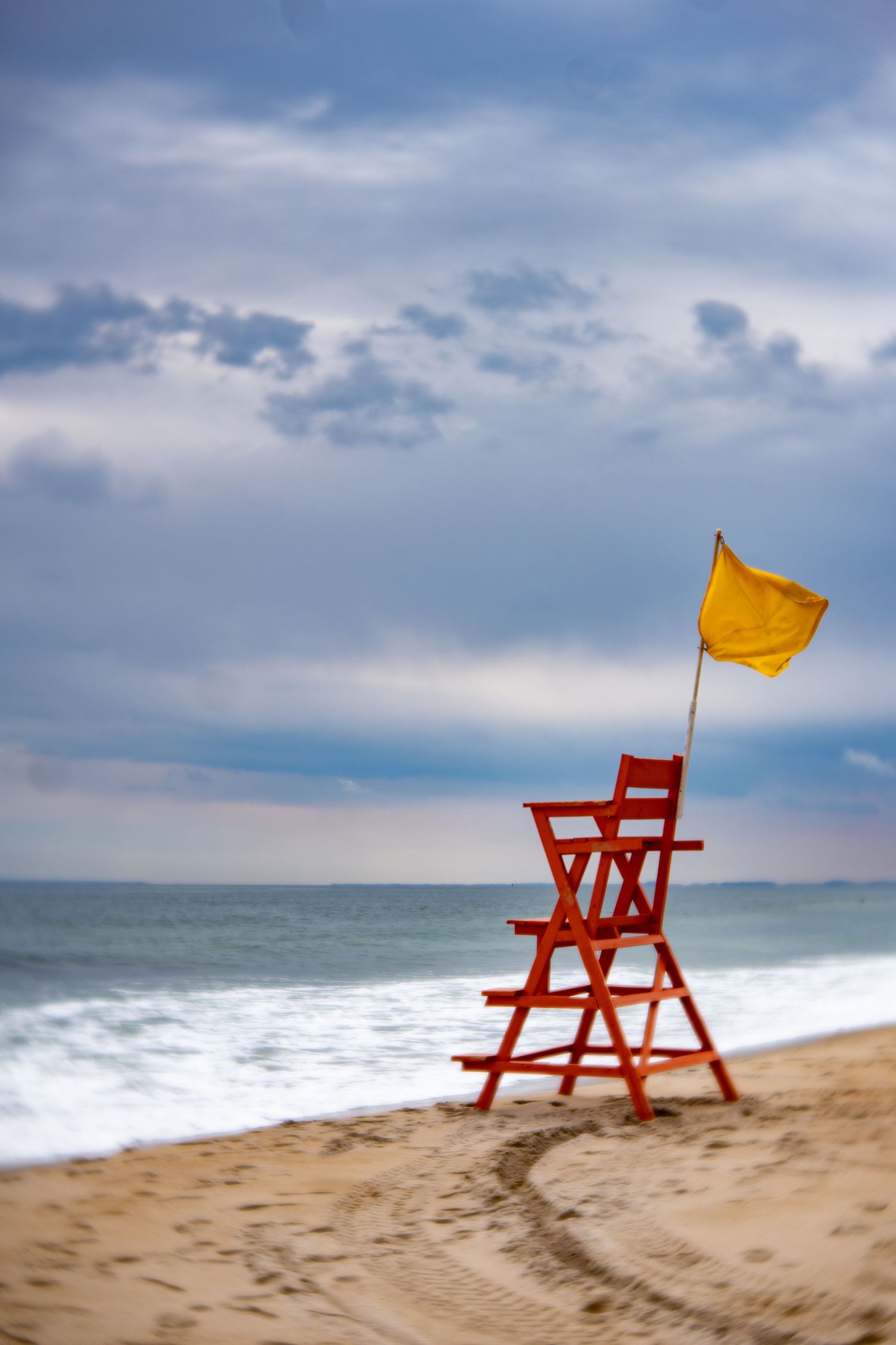 Red lifeguard chair with yellow warning flag on sandy beach under cloudy sky near ocean waves.
