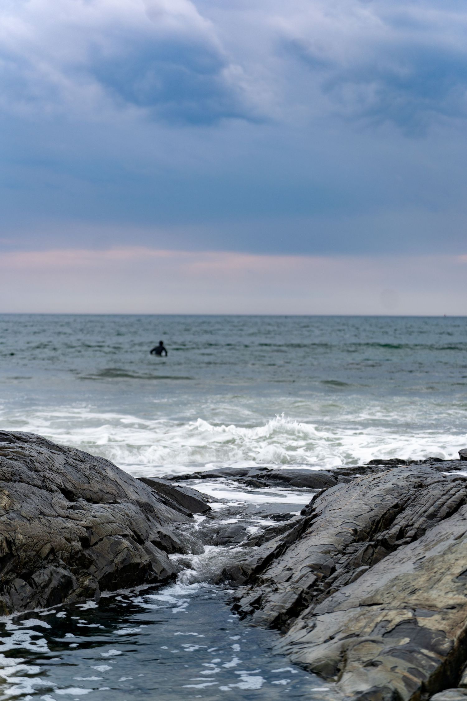 A lone surfer in the ocean waves with rocky foreground and dramatic cloudy sky at a rugged coastline.
