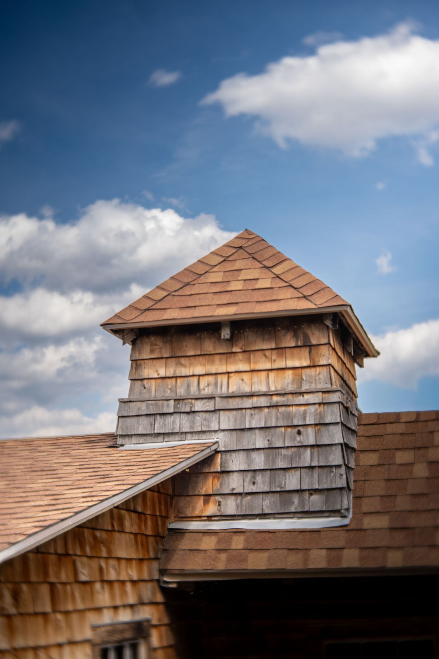 Rustic wooden cedar shake roof with cupola tower against a blue sky with white clouds.