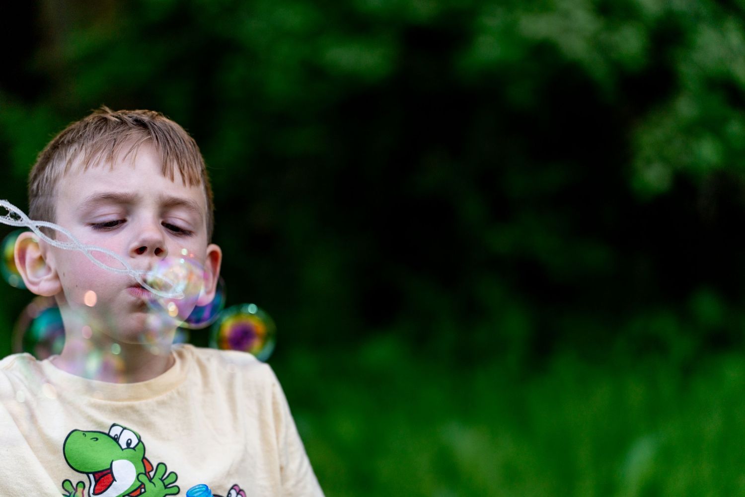 A young boy blowing colorful soap bubbles outdoors with green trees in the background.