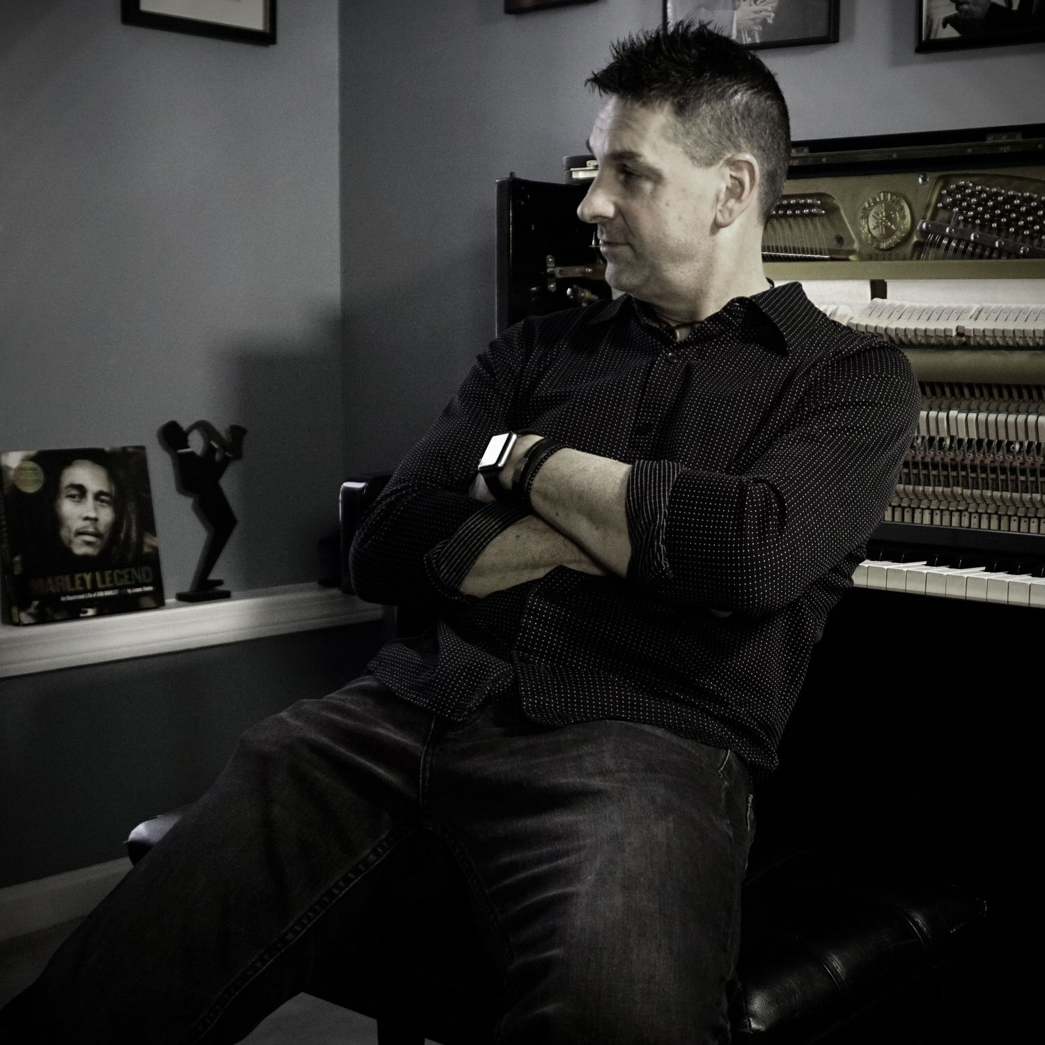 A dramatic black and white photo showing someone seated in front of a piano in a dimly lit room with framed artwork.