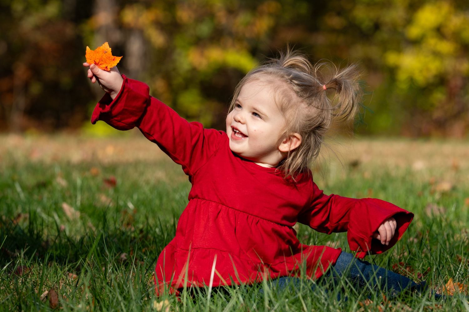 A child in a bright red sweater plays joyfully in autumn leaves on a grassy lawn.