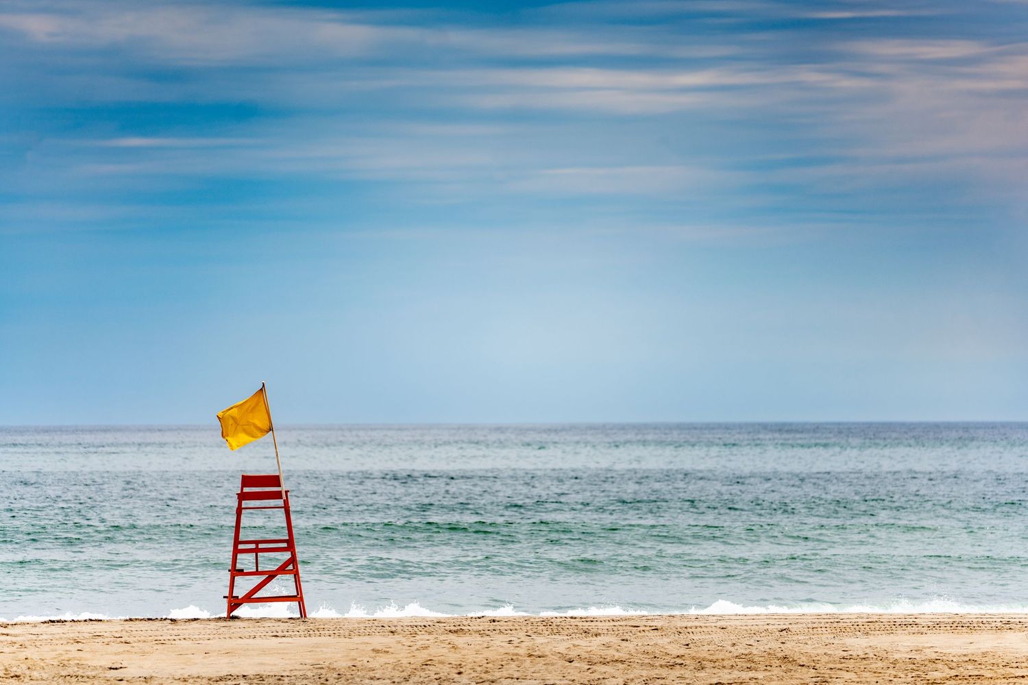 Red lifeguard chair with yellow flag on empty sandy beach with calm blue ocean and partly cloudy sky.