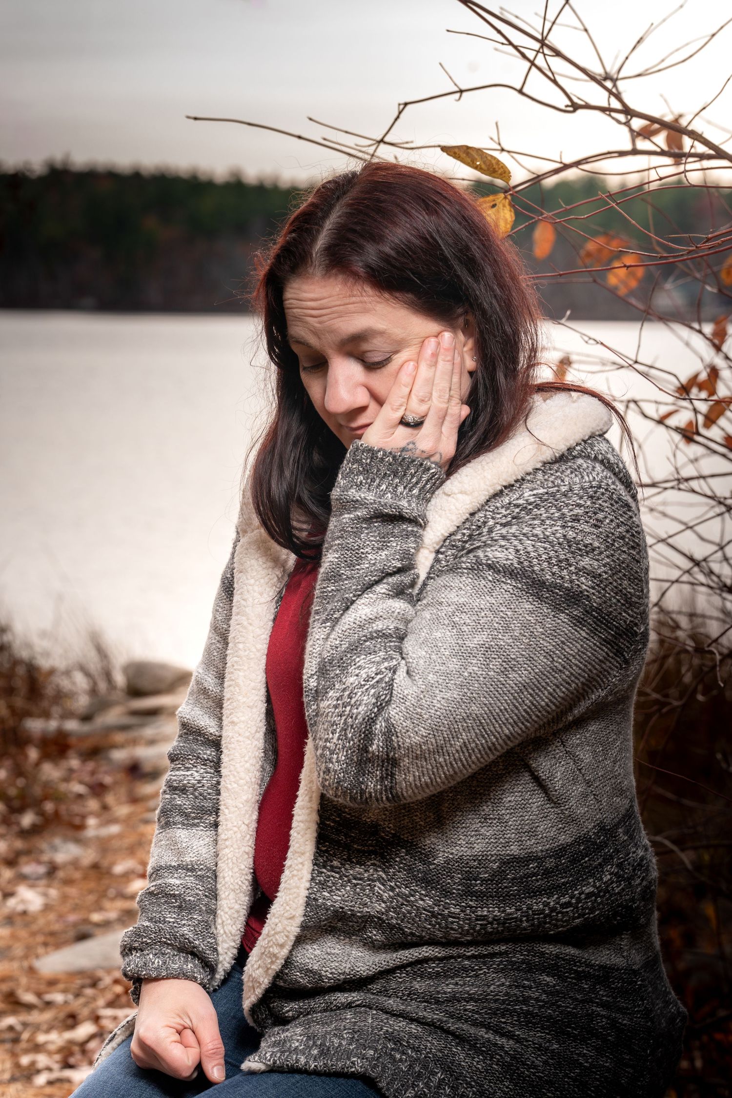 A person in a gray sweater sits outdoors among autumn branches with a burgundy scarf against a natural backdrop.