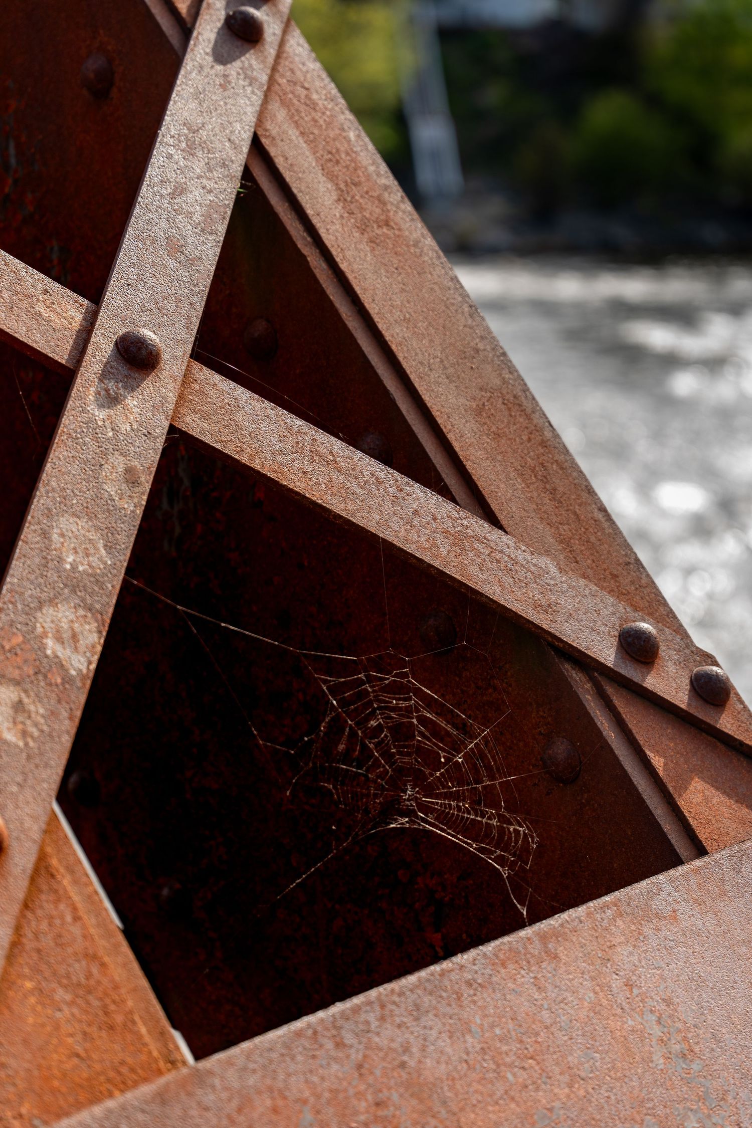 Rusty metal structure with a spider web in the corner, showing aged iron beams with bolts in natural light.
