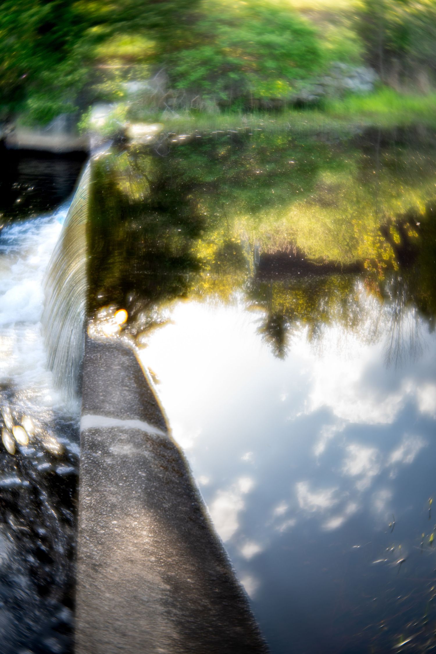 Blurred artistic photo of a small waterfall and dam with lush green trees reflecting in calm water below.