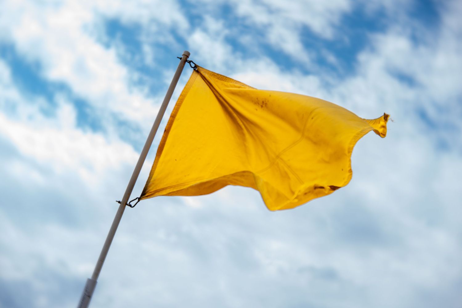 Yellow flag waving on a pole against a partly cloudy blue sky, symbolizing caution at a beach.