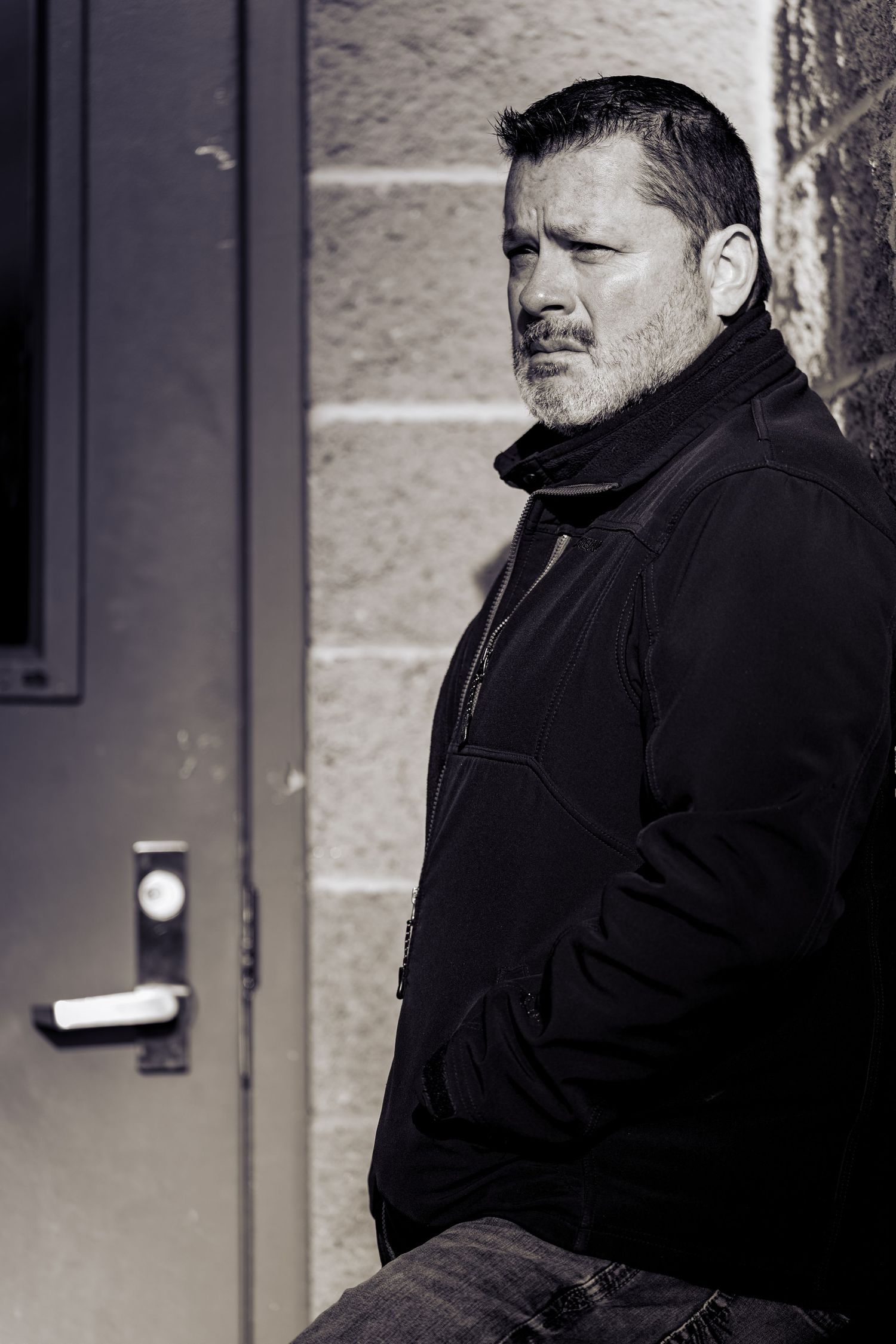 Black and white portrait of a middle-aged bearded man in black jacket leaning against a stone wall near a door.