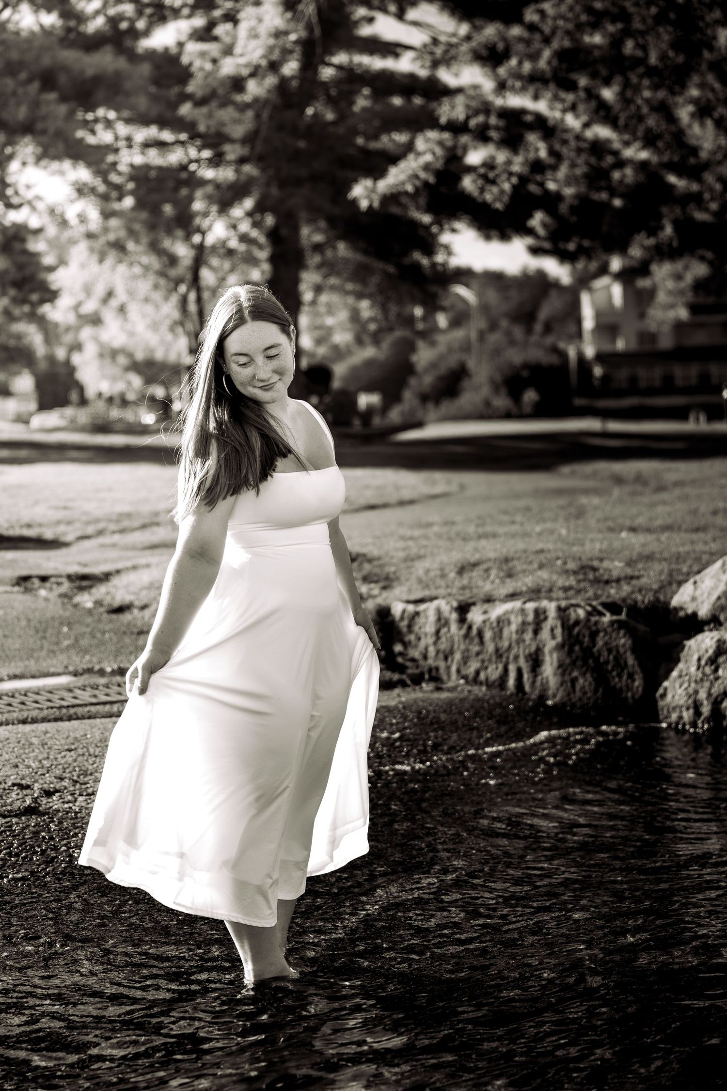 Black and white photo of a woman in a white dress wading barefoot in a stream in a lush park.
