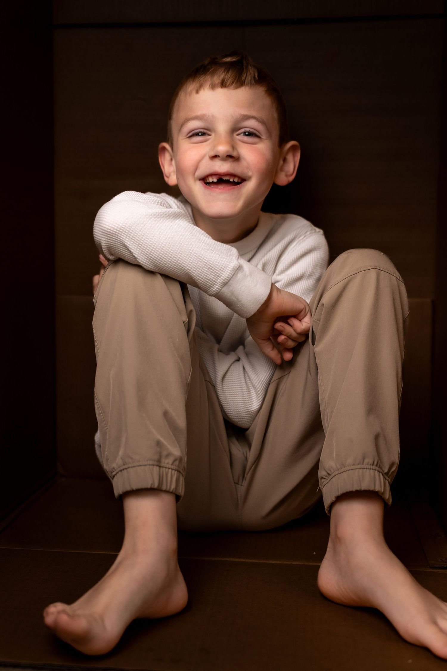 A barefoot young boy with a gap-toothed smile sits cross-legged on a dark floor wearing beige pants and a white sweater.