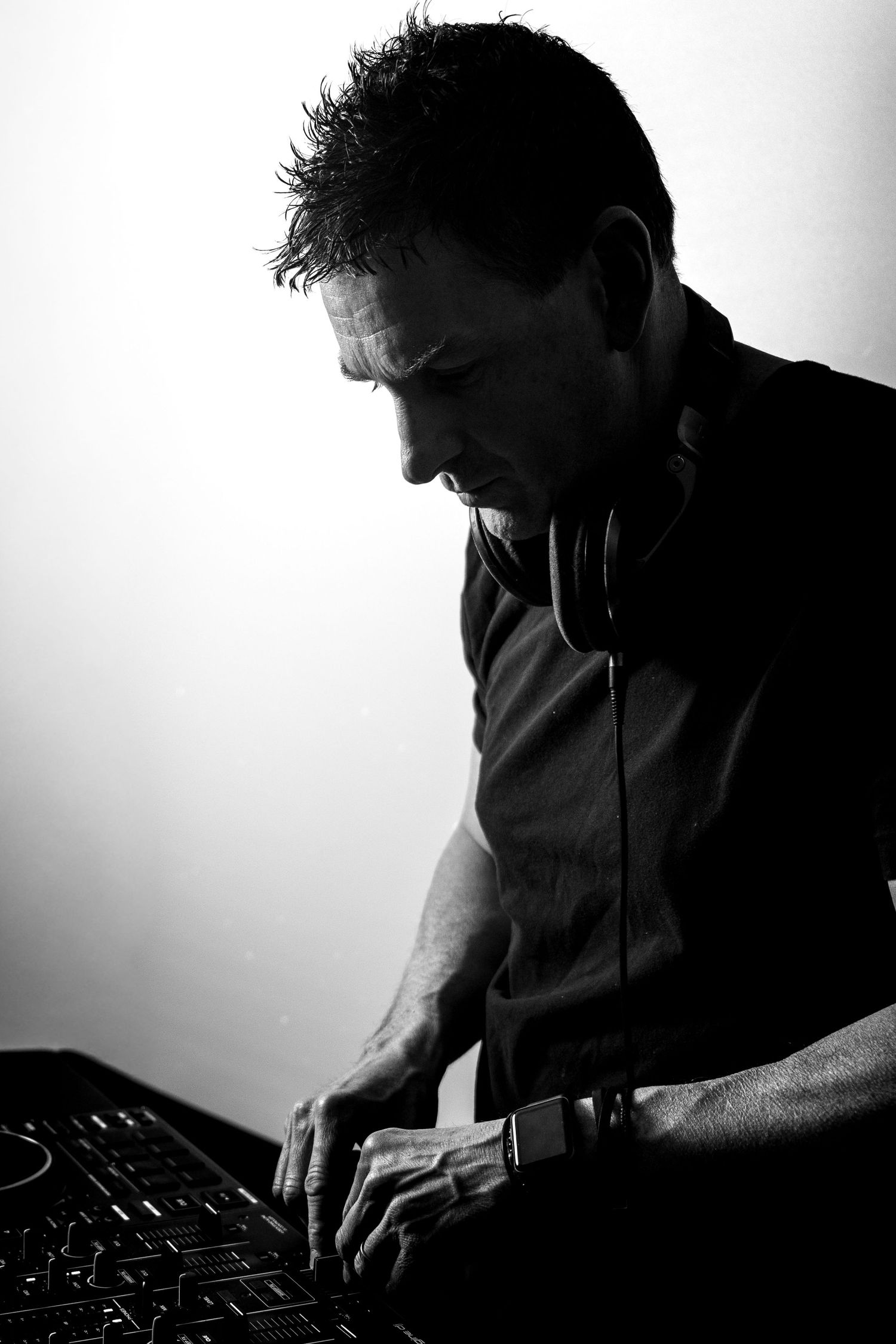Black and white photo of a male DJ wearing headphones around his neck, leaning over a mixing console.