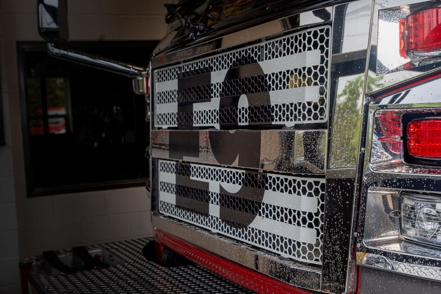 Close-up of a fire truck's chrome grille panel with bold lettering, parked inside a fire station bay.