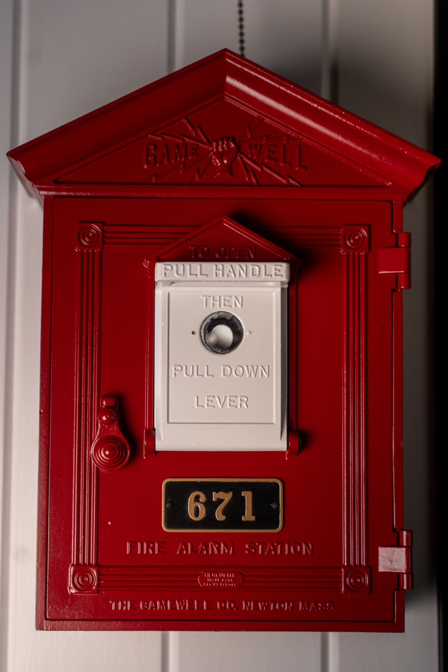 Vintage red fire alarm station box number 671 with pull handle lever mounted on a white wall.