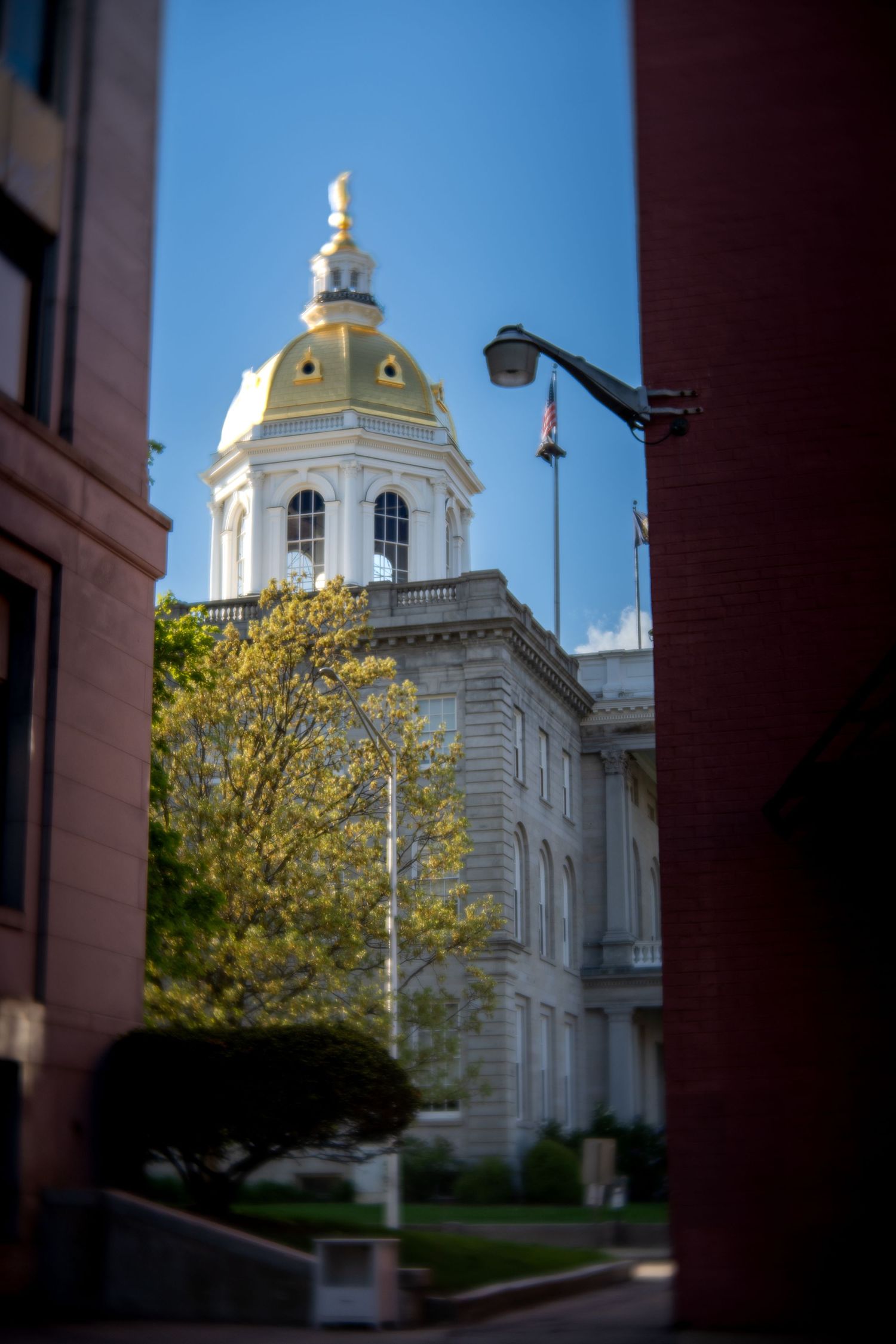 New Hampshire State House golden dome visible between brick buildings with American flag on blue sky day.