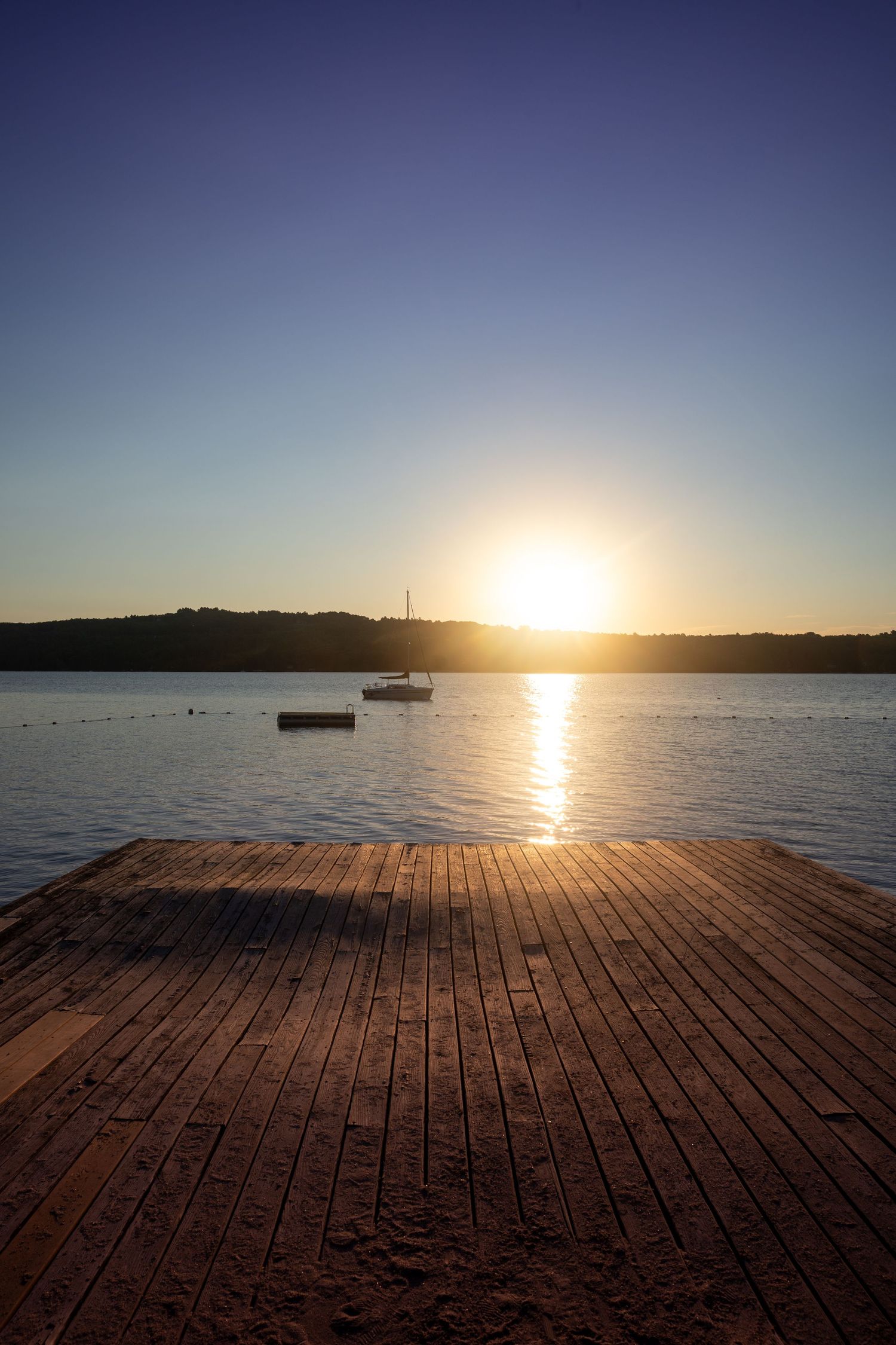 Panoramic sunset view over calm waters with wooden dock extending into lake and silhouetted boat in distance.