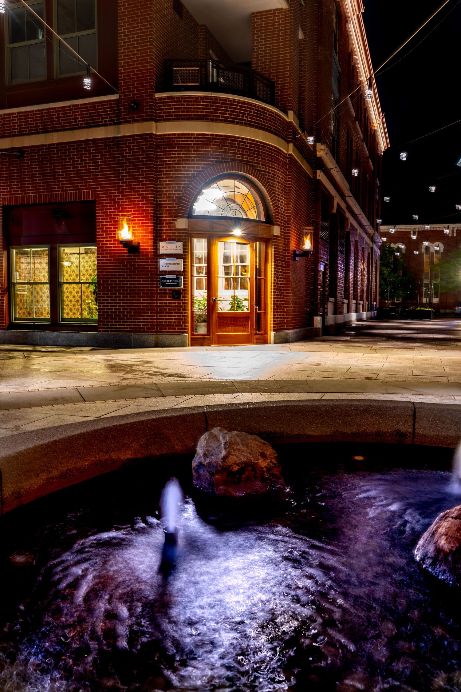 A brick building entrance with warm lighting reflects in a decorative outdoor fountain at night.
