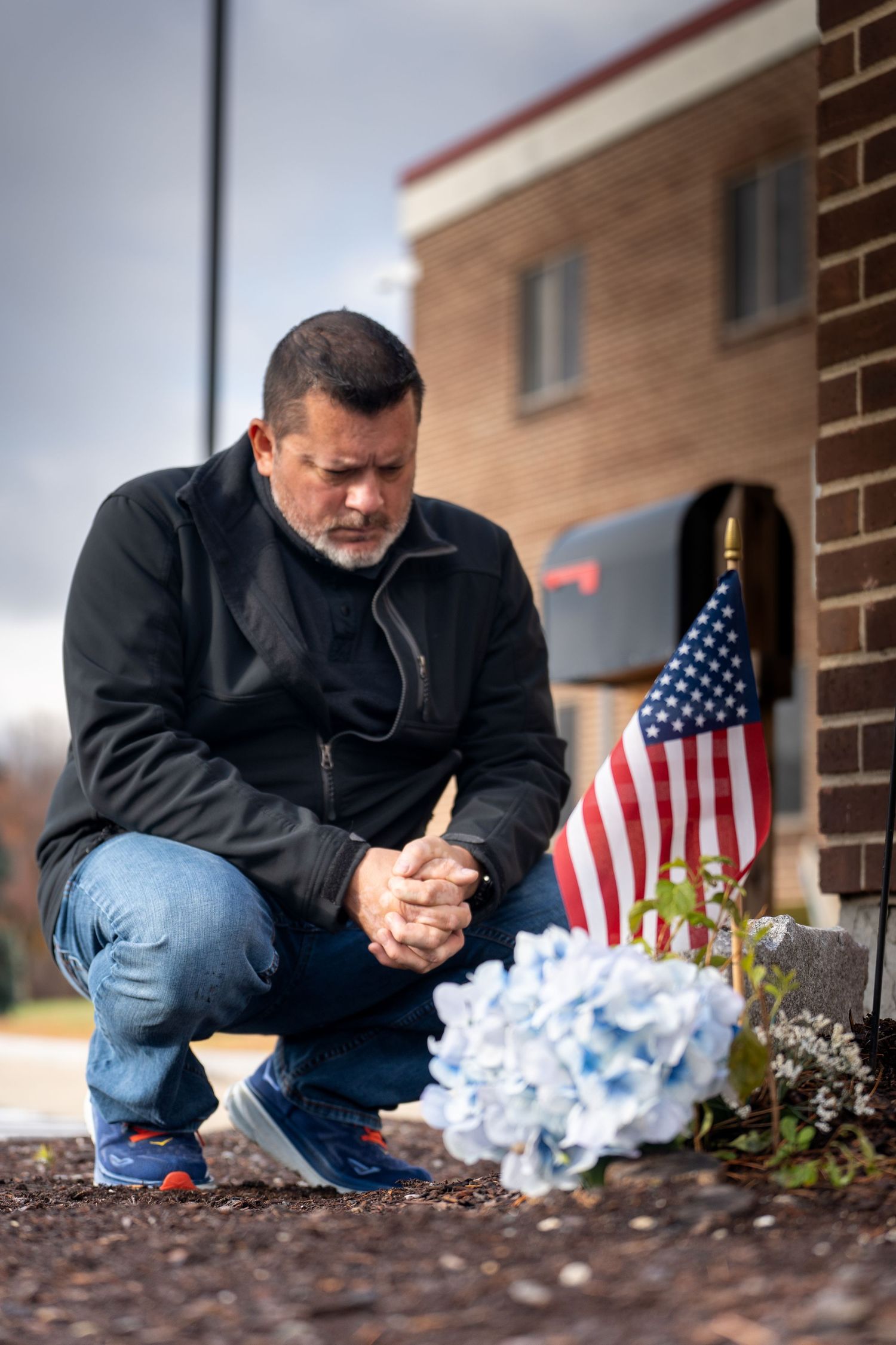 A person kneels in prayer beside American flags and white flowers outside a brick building.