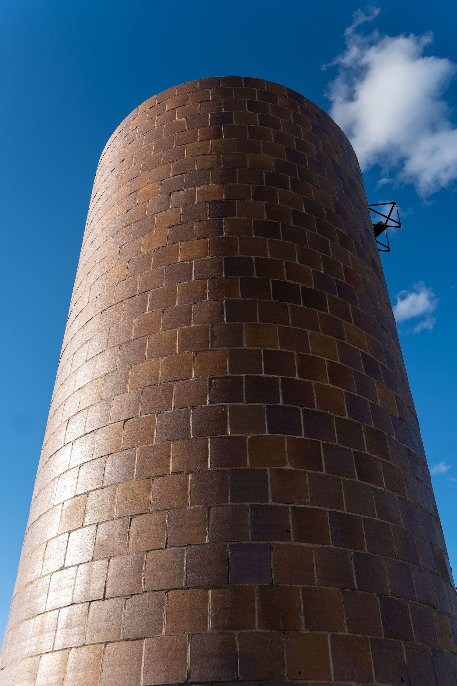 Tall cylindrical brick tower against a bright blue sky with white clouds, featuring a metal bracket near the top.