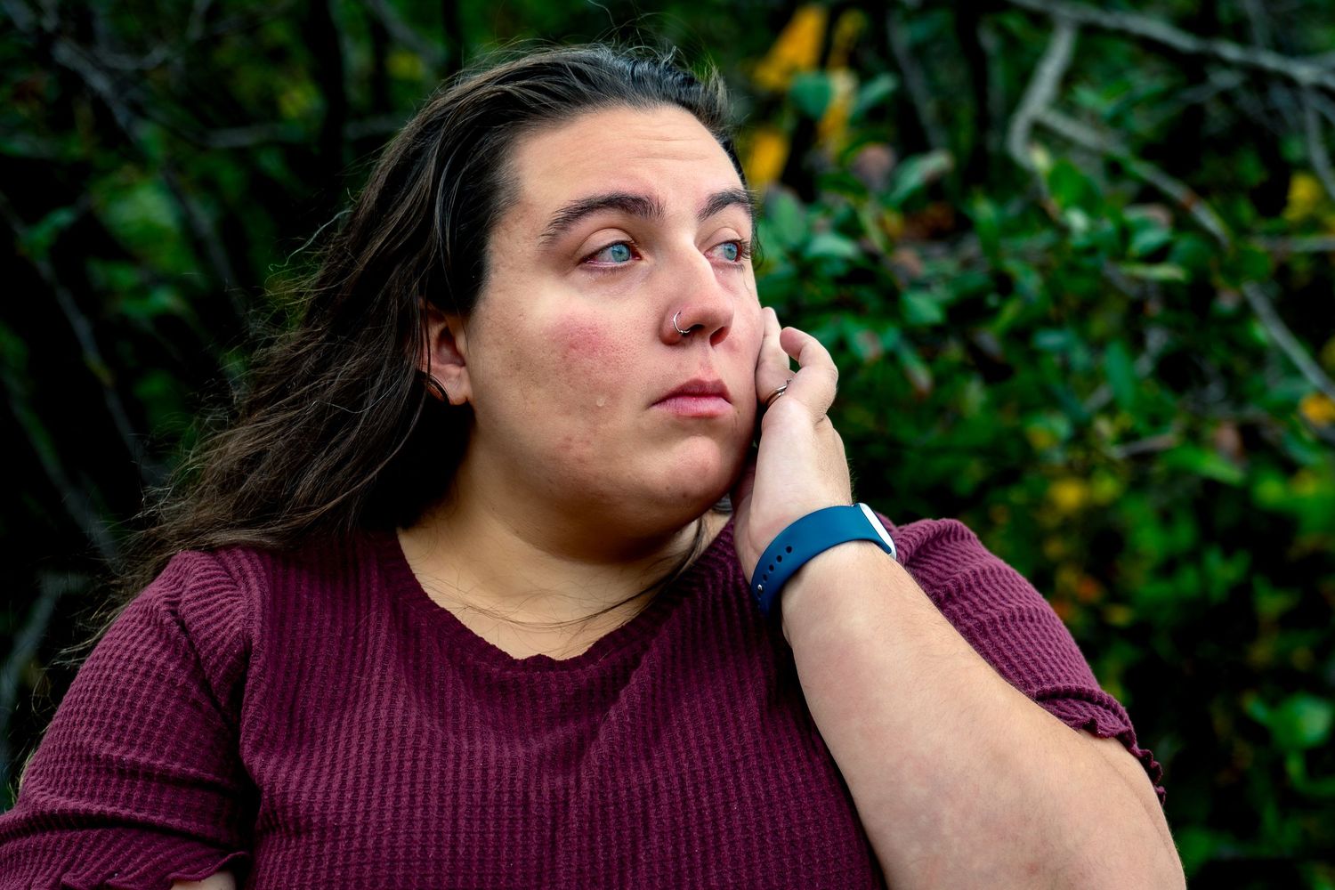 A person in a burgundy sweater sits thoughtfully with hand on chin against a green foliage background.