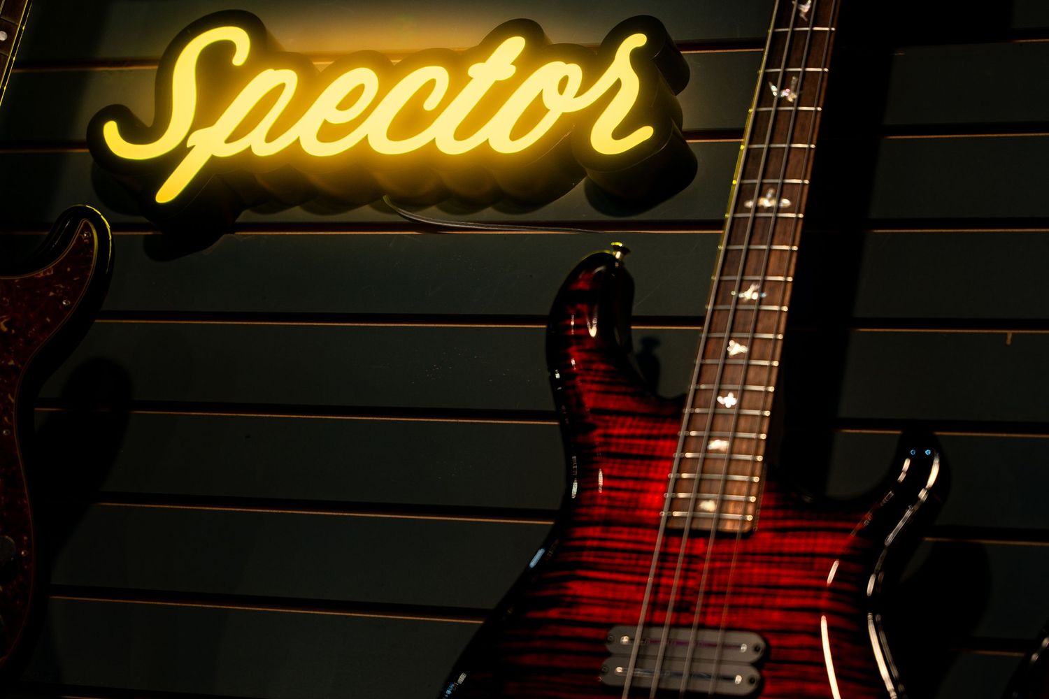 Red flame-top Spector bass guitar displayed under a yellow neon Spector brand sign.