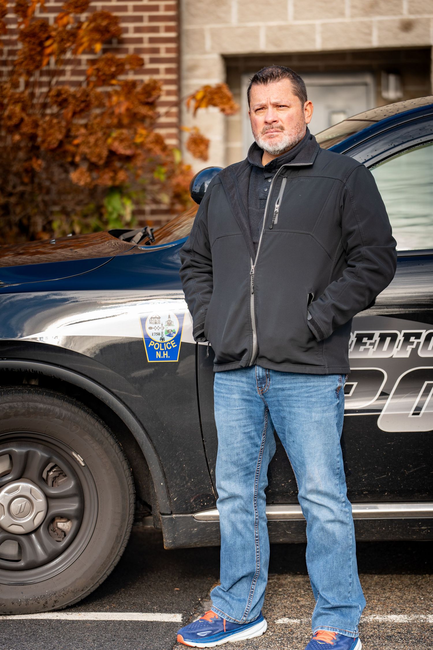 Person in black jacket and jeans standing beside police vehicles in a parking lot during autumn.
