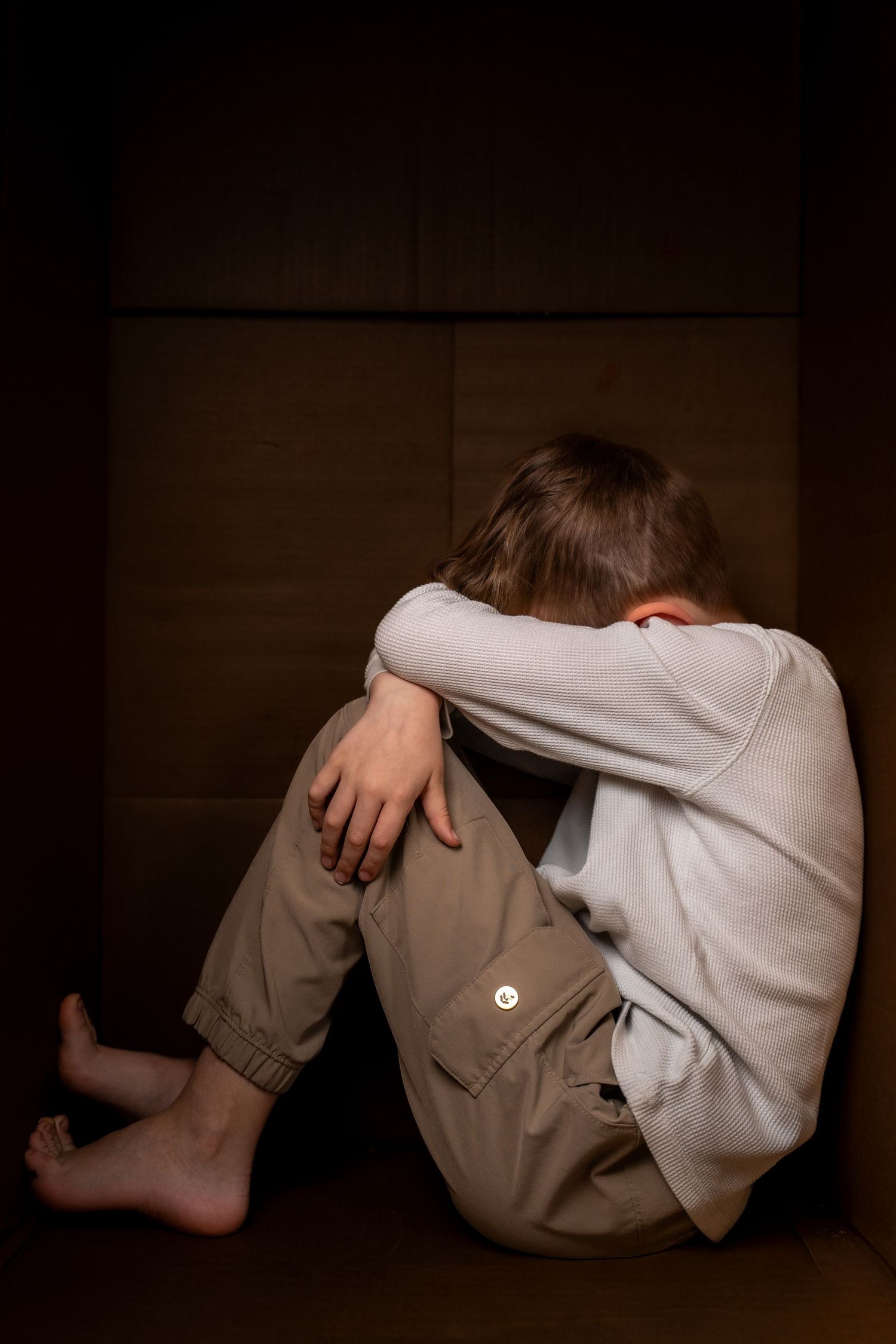 A sad barefoot child sits alone in a dark corner, hugging their knees with head down, wearing beige pants and gray shirt.