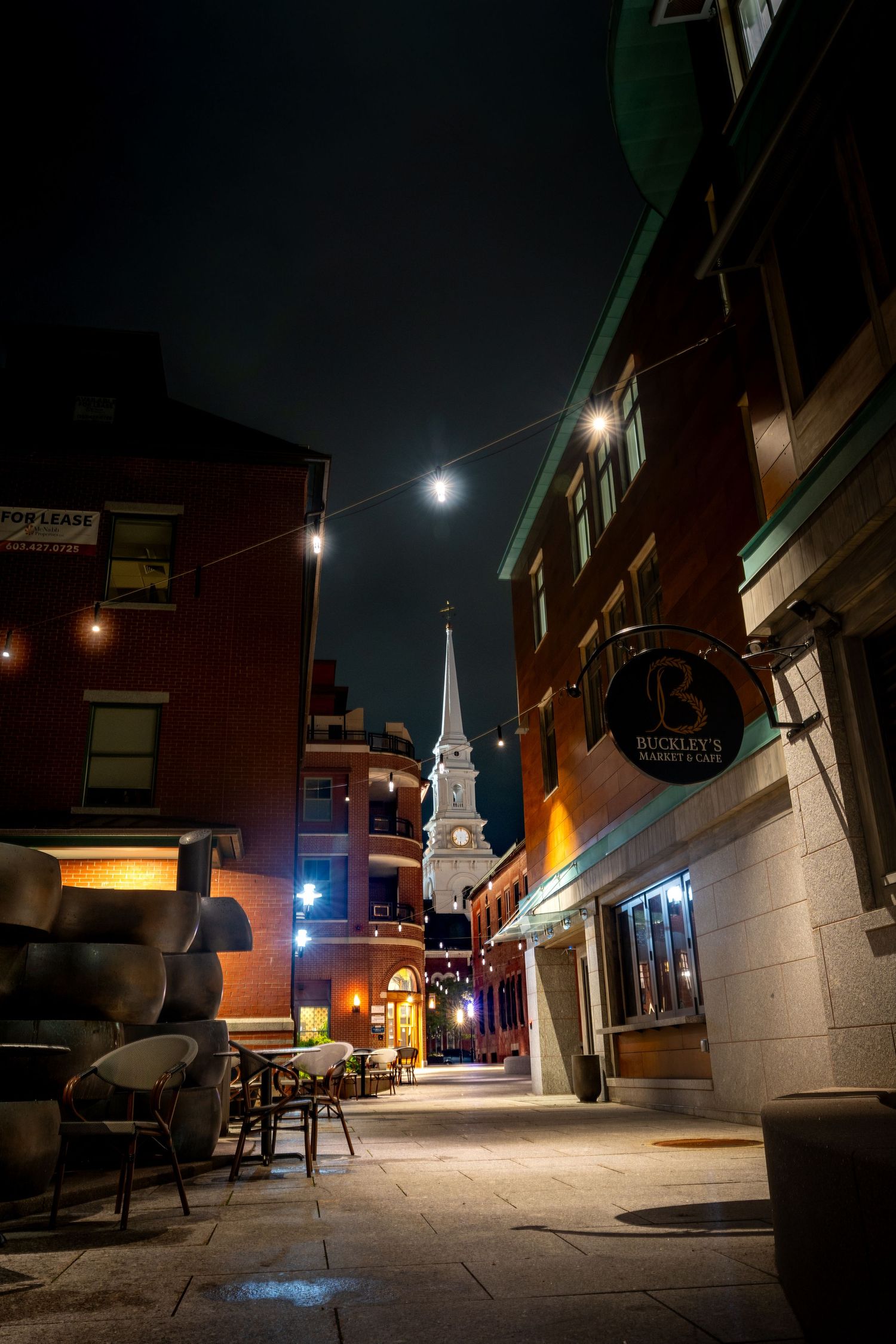 A moonlit city alley at night with string lights, brick buildings, and a white church steeple in the background.