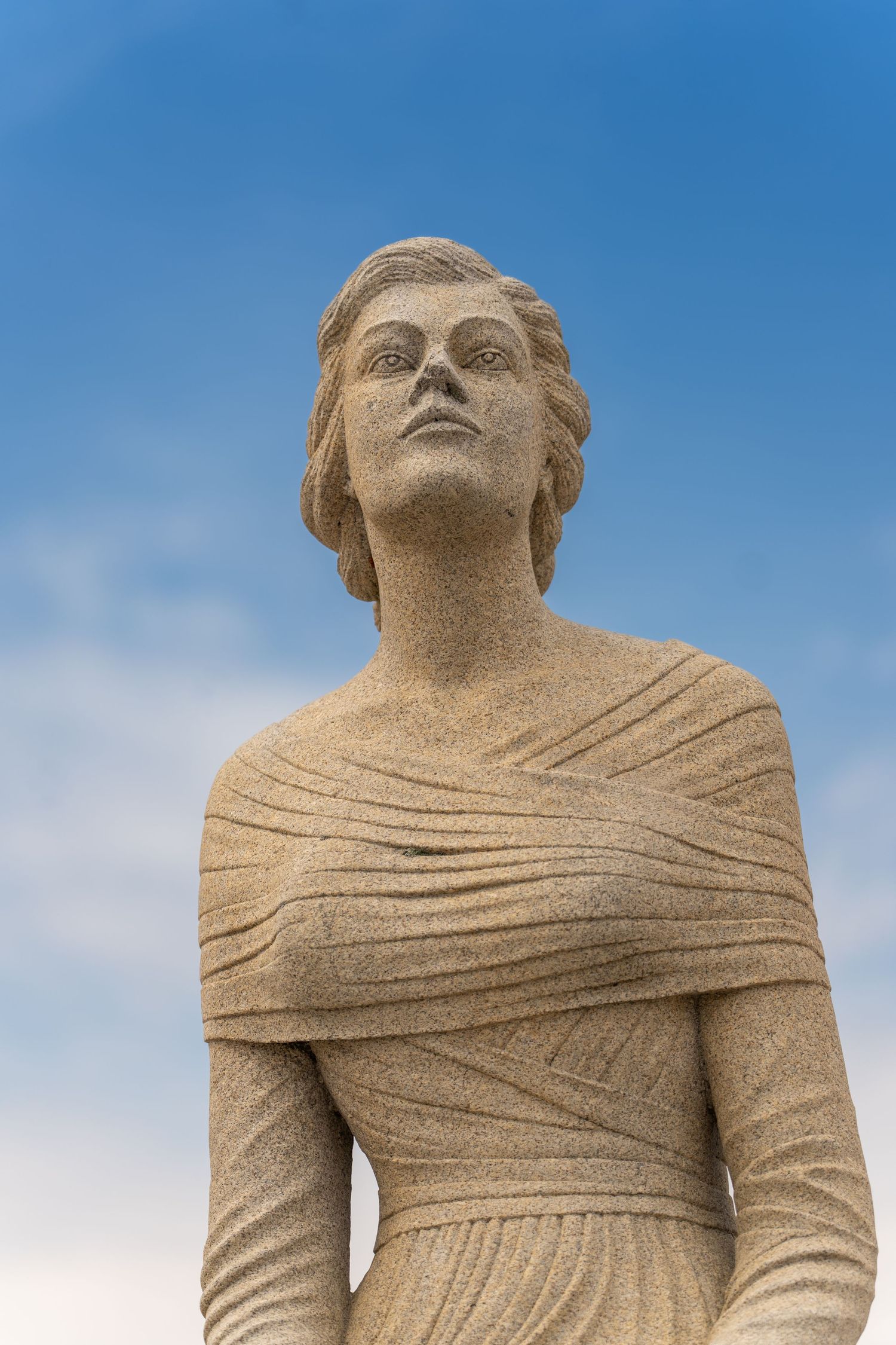 Stone statue of a woman gazing upward against a blue sky, detailed granite sculpture with draped clothing.