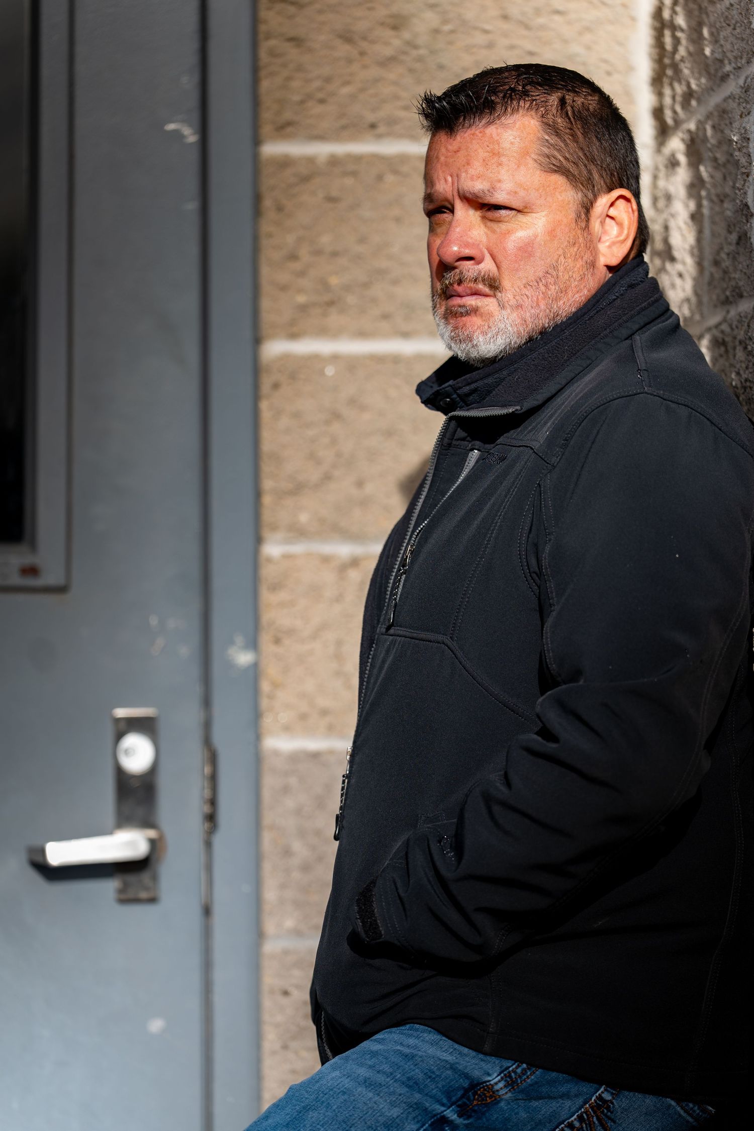 Person wearing a dark jacket sits alone against a concrete wall looking downcast.