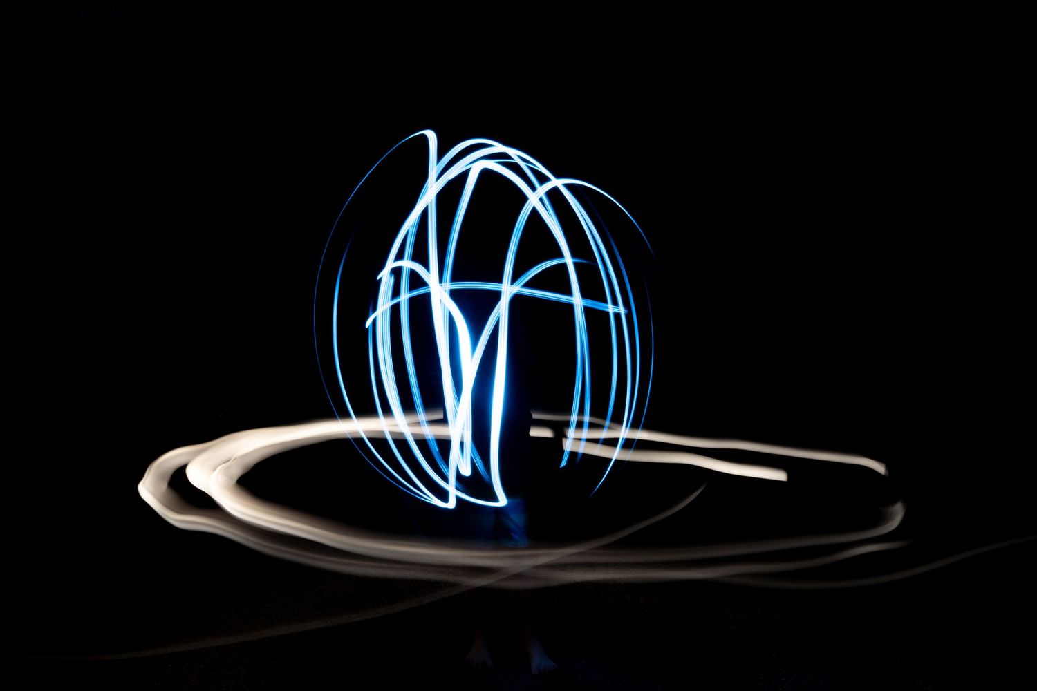 Long exposure light painting photography showing glowing blue and white light trails forming a sphere on black background.