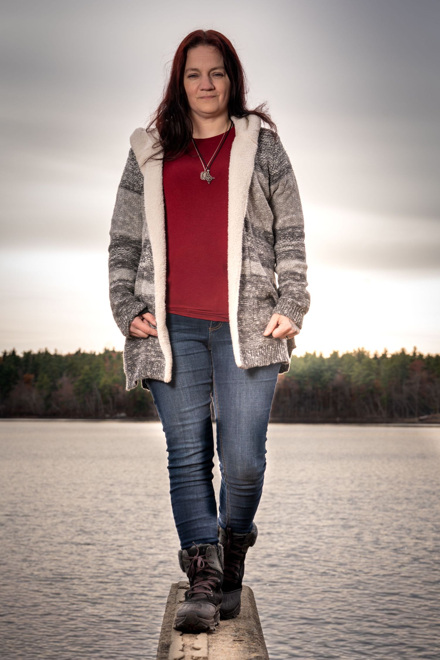 Person wearing red top and gray patterned jacket poses by serene lakeside at sunset in casual outdoor photoshoot.