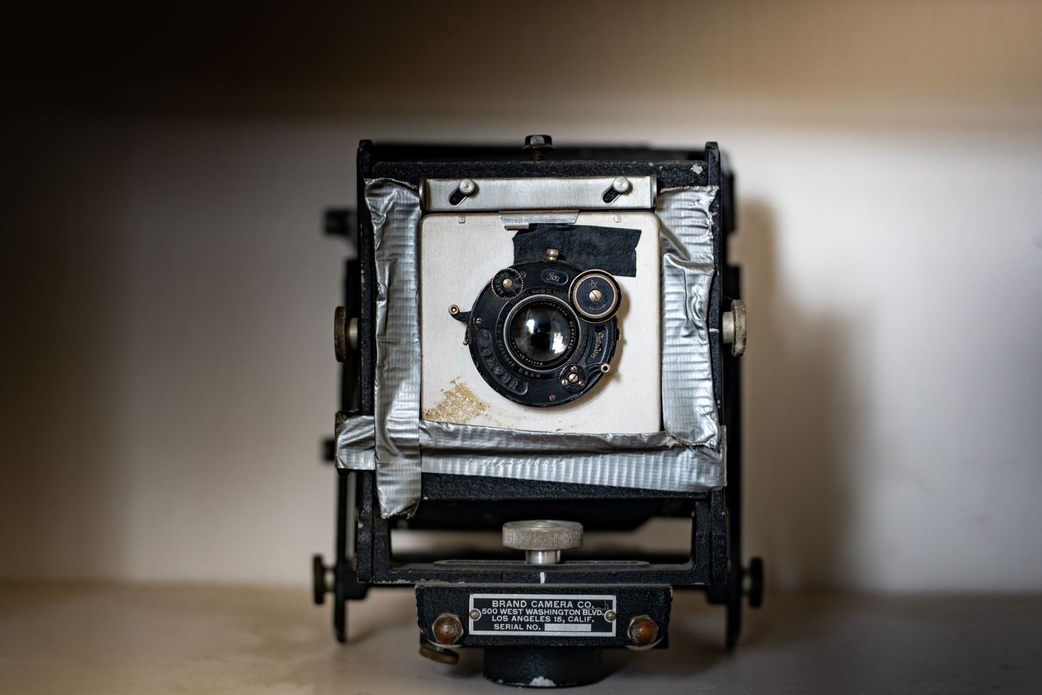 Vintage large format film camera with bellows and lens board, wrapped in silver tape, displayed on a light background.