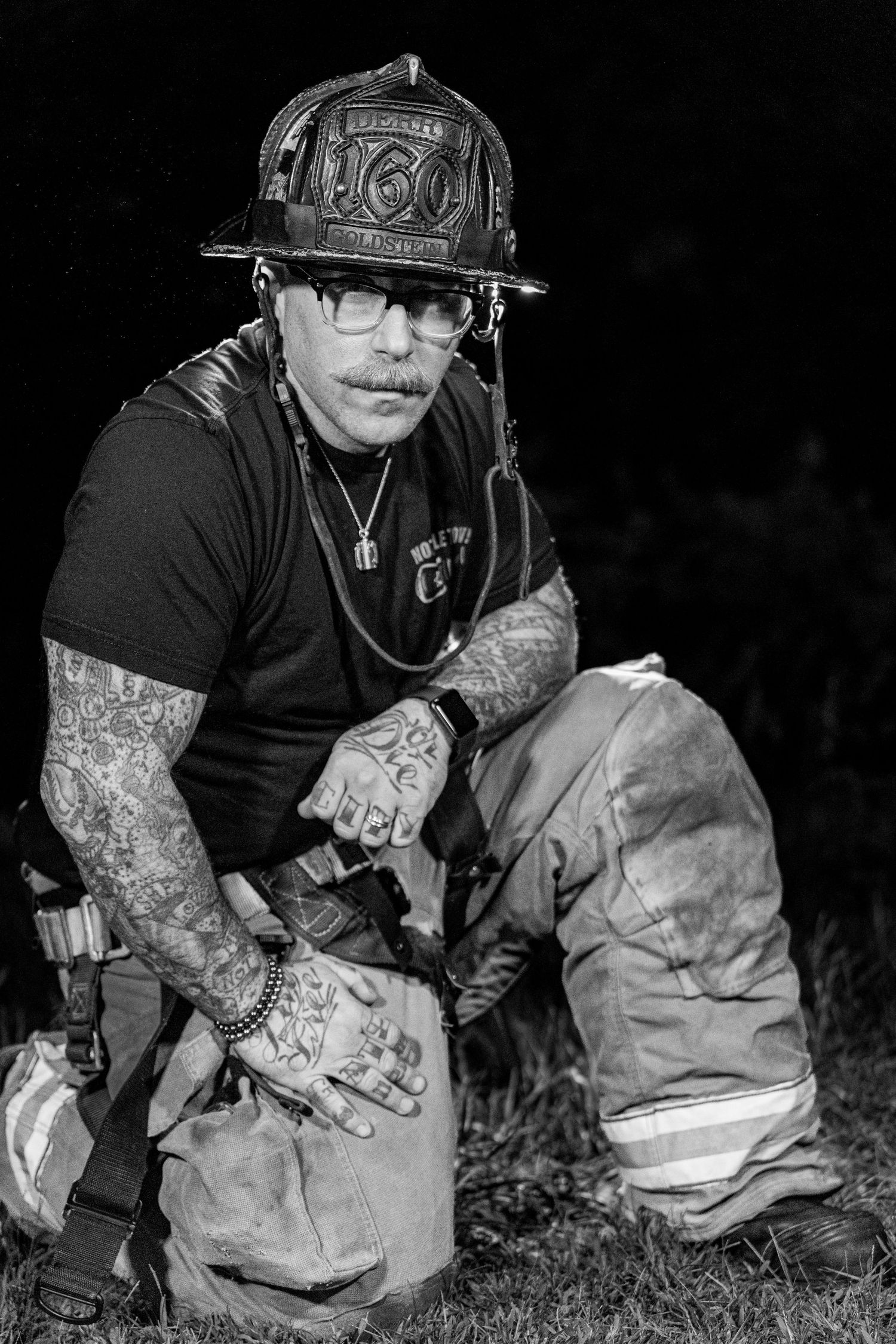 A firefighter in protective gear with visible tattoos kneels down during an emergency response operation.